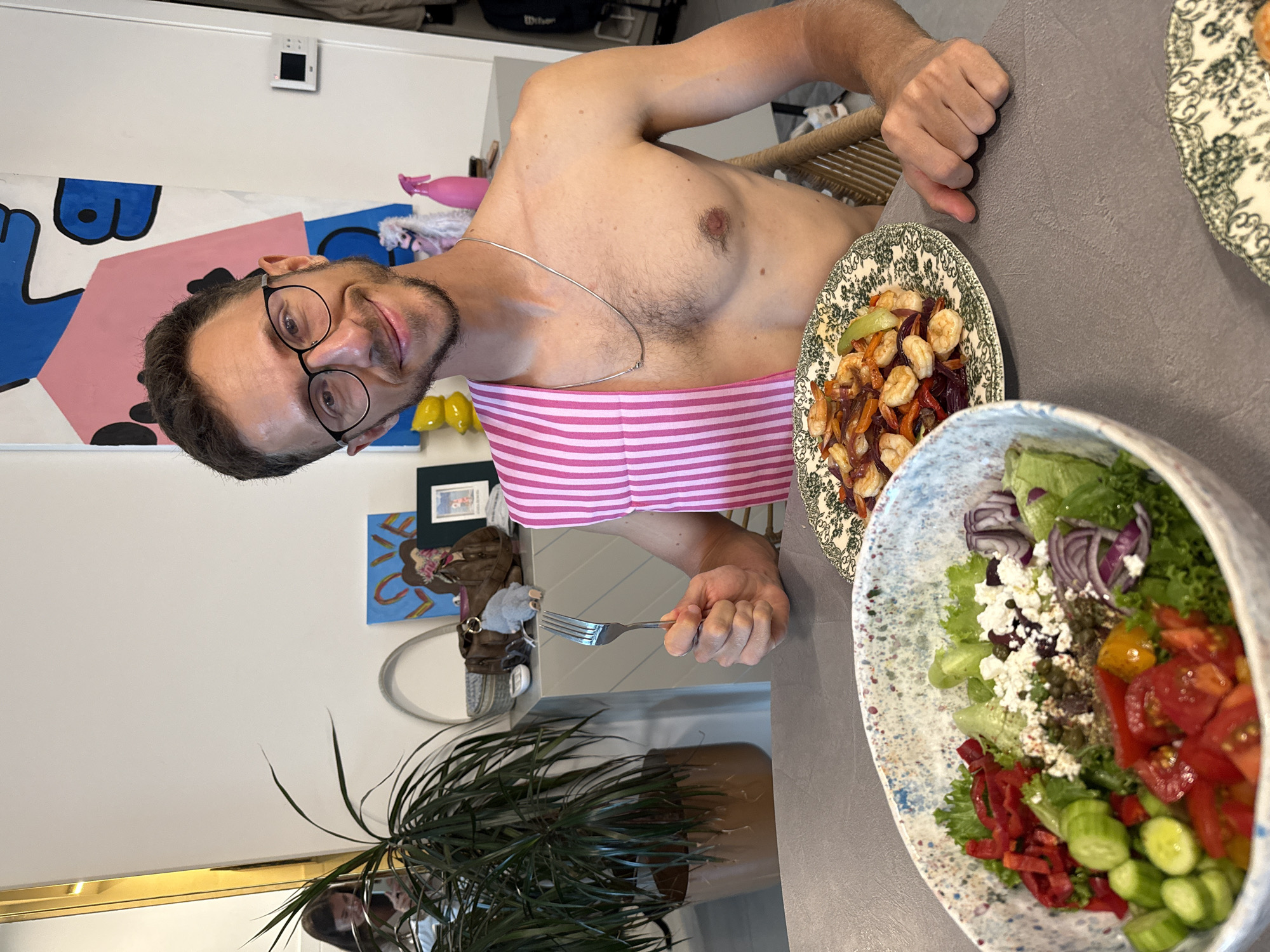 A man wearing glasses and a pink and white striped top sits at a gray table with two bowls of food. One bowl contains a salad with tomatoes, lettuce, cucumbers, feta cheese, and red onion. The other bowl appears to have shrimp and vegetables. The man holds a fork in his left hand. A potted plant is visible in the foreground. Behind the man are various decorations on a white wall. The city and specific location are unknown.