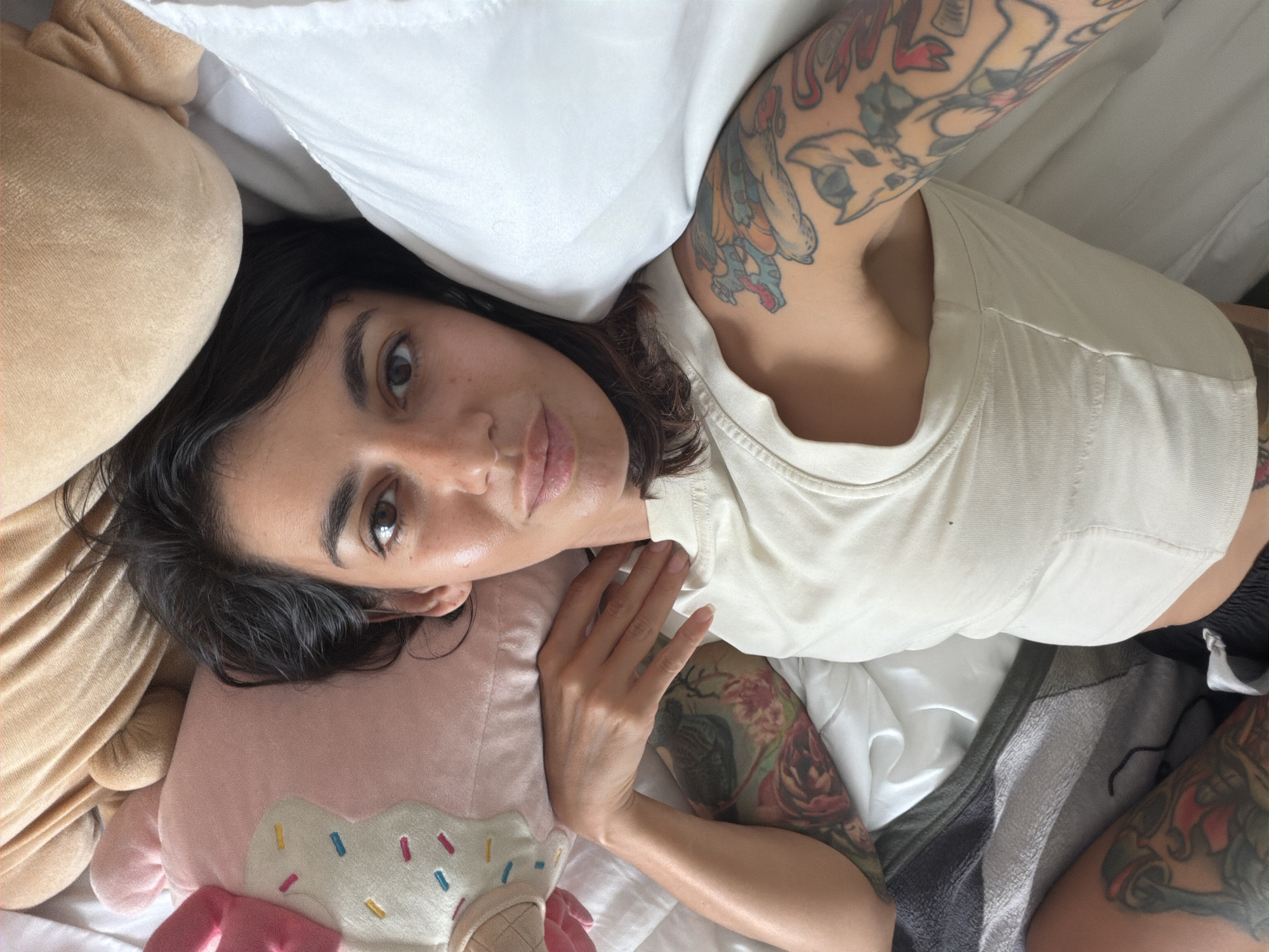 A female individual is depicted lying on her back, looking directly at the camera. She has a medium complexion, dark hair, and brown eyes. Her lips are slightly parted. Visible tattoos cover her left arm and shoulder, depicting stylized animals and other figures in vibrant colors, including a blue creature resembling a dragon or monster, and a cat-like figure. Additional tattoos with floral patterns are visible on her right arm and on her left thigh. She wears a cream-colored, ribbed sleeveless top. Her right hand is placed near her neck. She is surrounded by bedding and plush items; a large, light brown furry object is behind her head, a white pillow is to her right, and a pink, cupcake-shaped pillow with colorful sprinkles is to her left. White sheets cover the bed, and a grey and black striped blanket is partially visible in the lower left. The scene is indoors, likely a bedroom, with bright, diffused lighting. The location, according to the provided information, is Kuta Selatan, Indonesia.