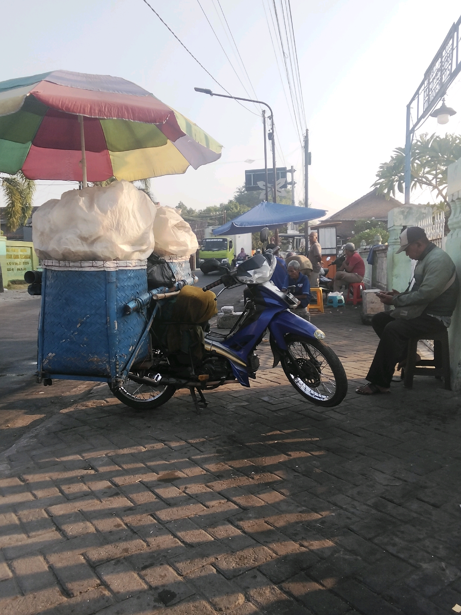 The image captures a street scene in Singogalih, Indonesia, at what appears to be sunrise or sunset due to the long shadows cast on the brick-patterned sidewalk. In the foreground, a blue motorcycle is parked, equipped with a makeshift cargo carrier. This carrier, fashioned from a blue woven basket, is laden with two large, white, pillow-like sacks. An open, multi-colored umbrella (red, yellow, green, and white) is positioned above the sacks, providing shade.
To the right of the motorcycle, a man wearing a cap, a green jacket, and dark pants is seated on a dark wooden stool, engrossed in his mobile phone. His footwear appears to be sandals. Further back, several other individuals are visible, some seated on stools and others standing. One man in a red shirt is sitting on a red stool. Behind them, a blue umbrella is open, and a building with a pitched roof can be seen, along with trees and a street lamp. The street itself is paved with interlocking bricks. In the background, a green truck is parked. The overall atmosphere suggests a quiet moment in a roadside setting.