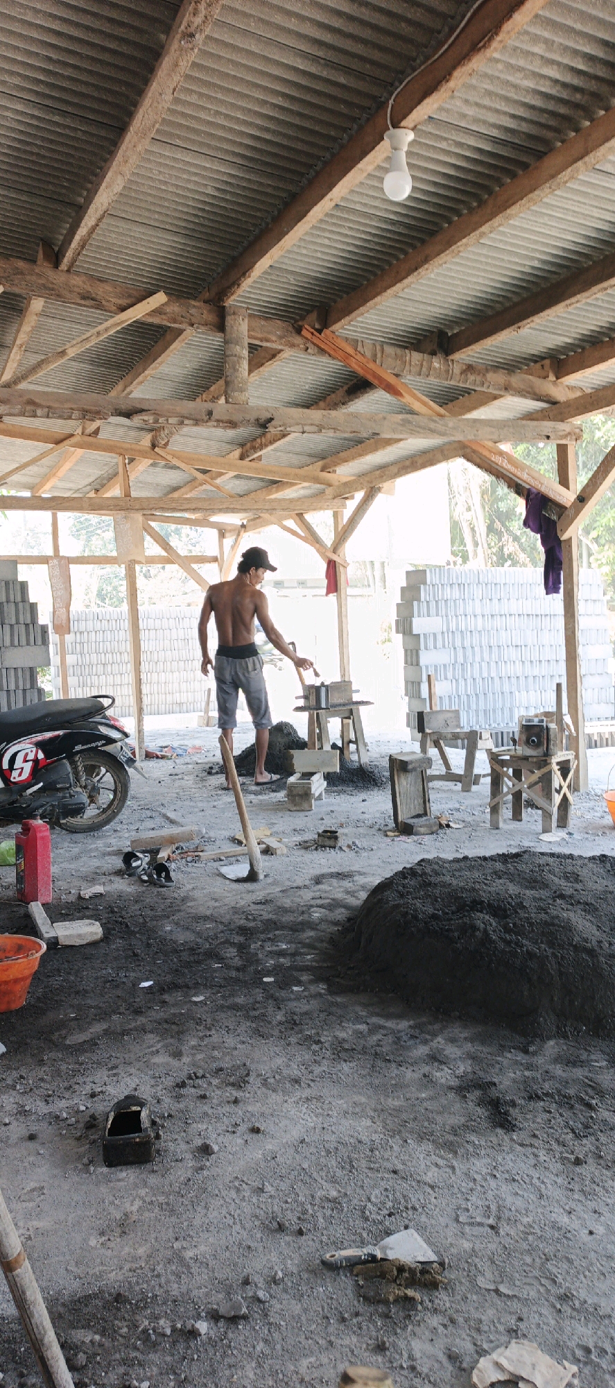 The image shows an outdoor construction site, likely in Kasiyan, Indonesia, where bricks are being manufactured. A shirtless man wearing gray shorts and a dark baseball cap is working at a table with a brick-making mold. He appears to be using a tool to press material into the mold. Various stacks of finished bricks are visible in the background, along with piles of raw materials and construction tools such as a shovel and buckets. A motorcycle is parked on the left side of the frame. The structure overhead, with a corrugated metal roof supported by wooden beams, suggests a rudimentary workshop or storage area. The ground is a mix of dirt and concrete, with scattered debris.