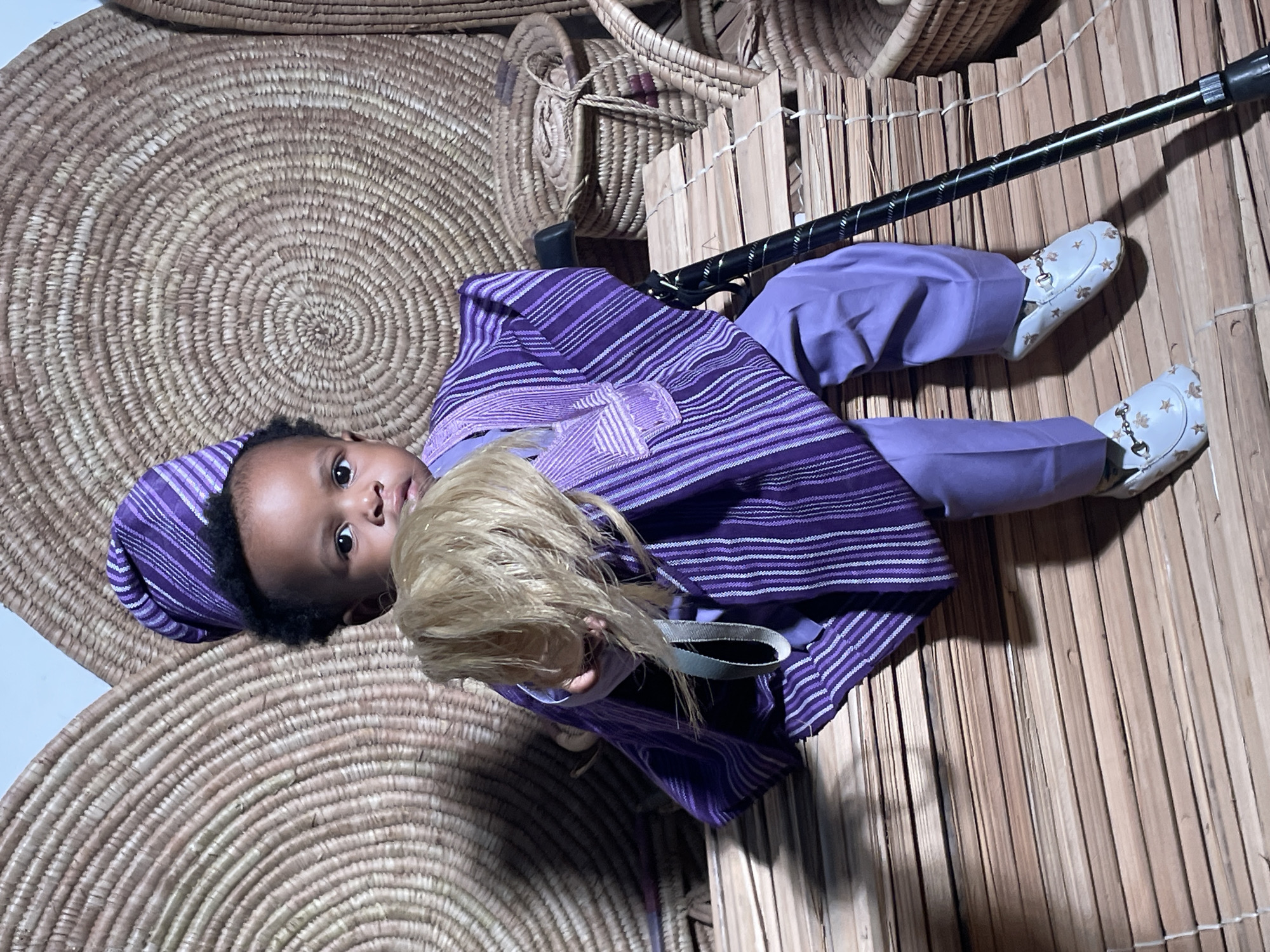 A child wearing a purple and white striped outfit, including a matching hat, is holding a doll with blonde hair. The child is lying on a woven bamboo mat with a walking stick beside them. In the background, there are woven baskets of varying sizes. The location is Jos, Nigeria.