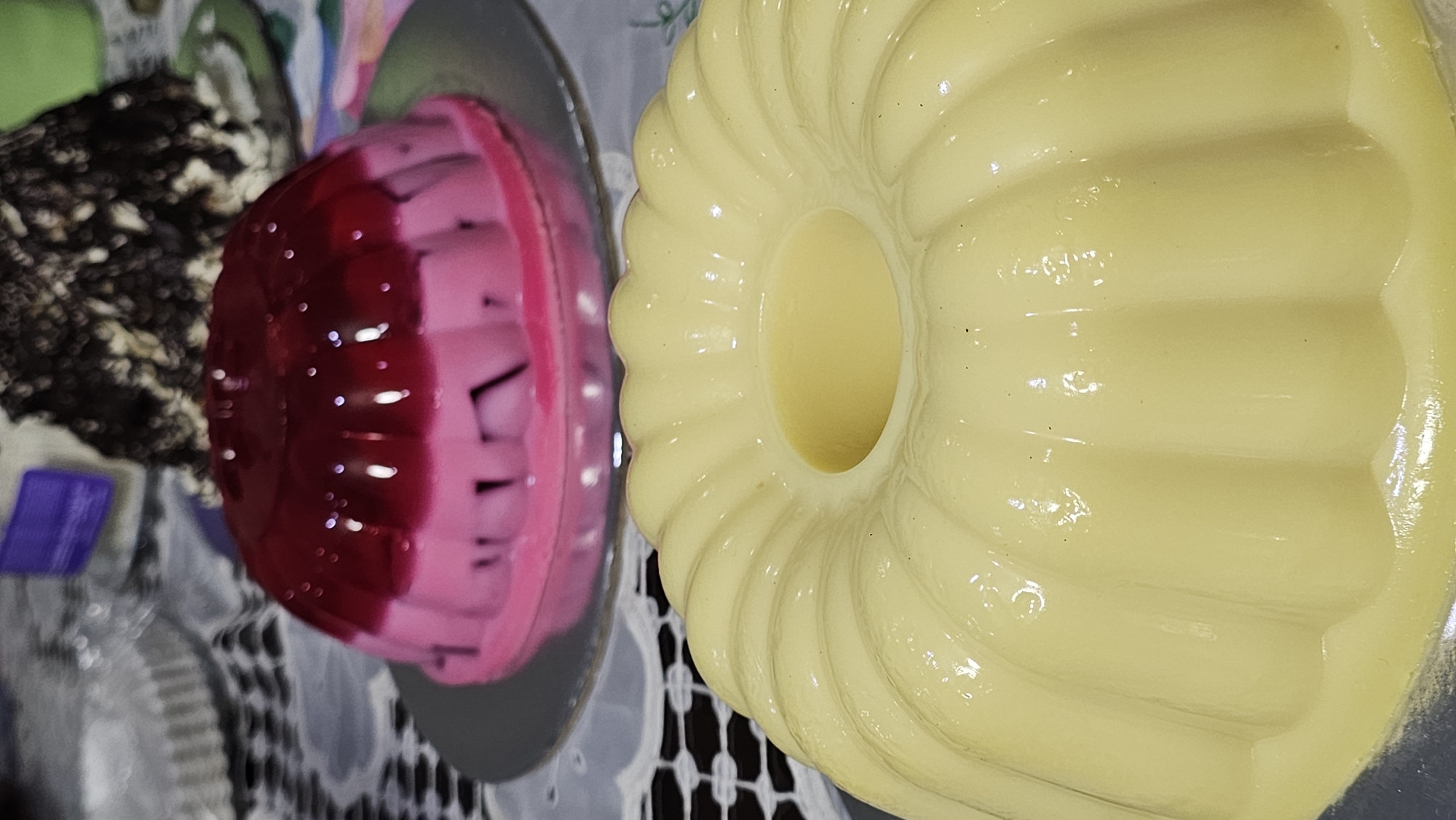 Two gelatin desserts are prominently displayed on a surface covered with a lace tablecloth. One dessert is yellow and molded in a ring shape with vertical ridges. The other is two-toned, with a red top layer and a pink bottom layer. It sits on a silver serving plate. Other objects are partially visible in the background, including a dessert with a dark, speckled appearance.
The location cannot be identified as San Bernardino Tlaxcalancingo, Mexico, from the visual information provided.