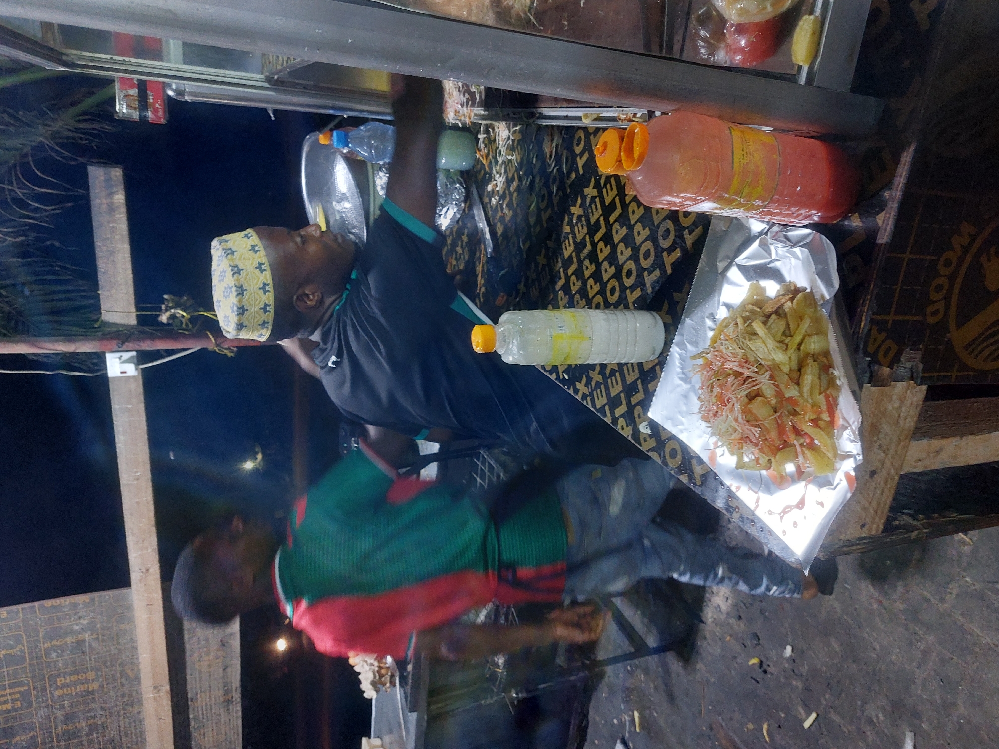 Here is a forensic description of the image:
The image depicts a street food vendor setting up his stall at night. The vendor is an adult male of African descent, wearing a yellow-patterned kufi cap and a dark-colored shirt with green trim. He is standing behind a metal food counter. A second adult male is to the left of the frame, wearing a red and green striped shirt.
On the counter, there are two large bottles of liquid, one orange and one clear. There is also a foil tray containing what appears to be french fries and shredded cabbage, potentially served with a reddish sauce. The counter surface is covered with a material printed with "TOPPLEX" and "WOOD DA" branding.
The setting appears to be outdoors at night, possibly in the city of Dar es-Salaam, Tanzania. The lighting is dim, likely from nearby streetlights. The background is dark and out of focus. A wooden board is visible along the bottom and left of the frame.
