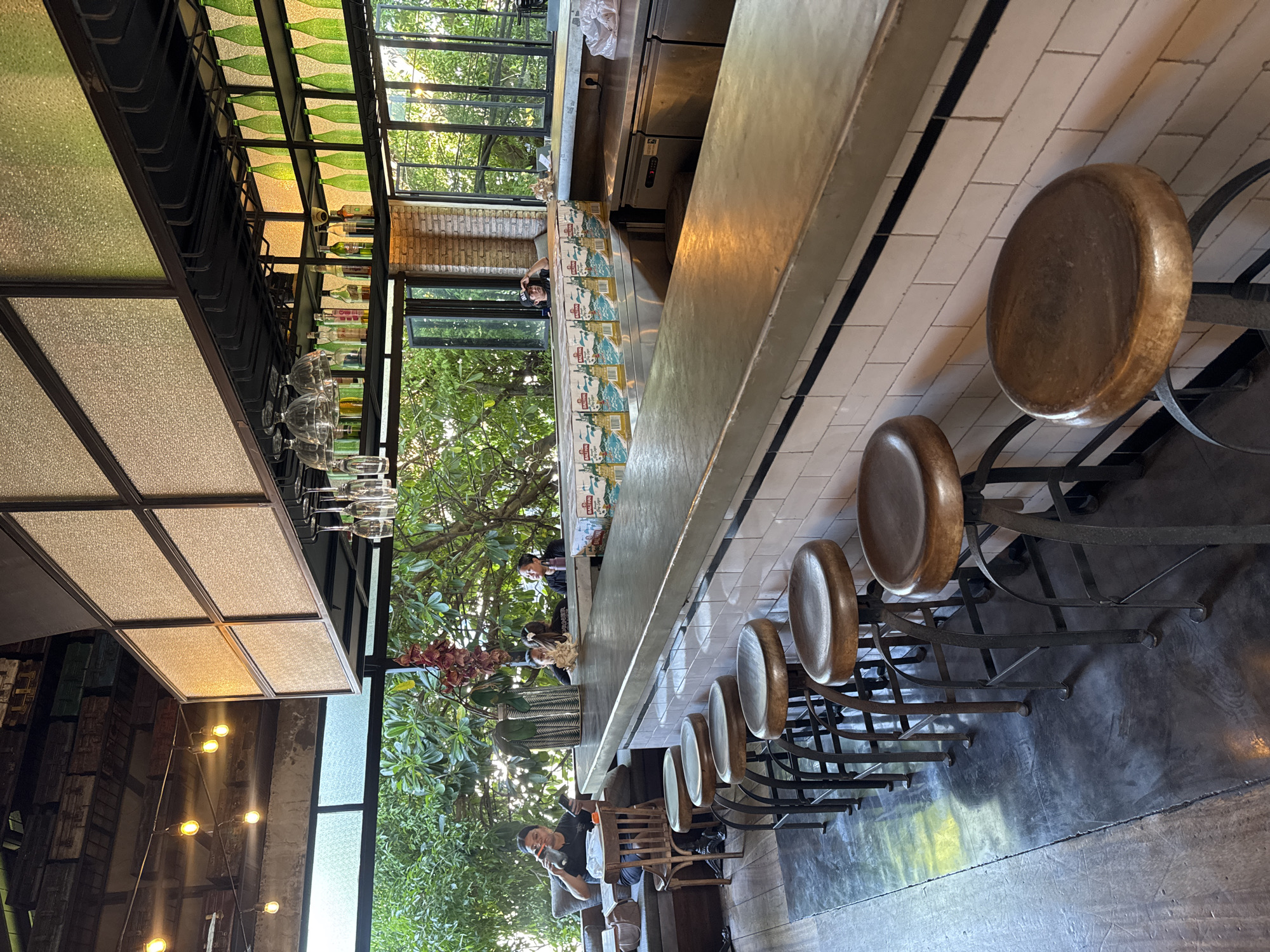 The image depicts the interior of a bar located in North Kuta, Indonesia, during daylight hours. The scene is dominated by a long, polished concrete bar counter with several wooden-topped bar stools arranged in a row. Behind the counter, a wall of white subway tiles runs along its base. Above the counter, a shelf displays numerous green glass bottles, some filled and others empty. Suspended from the ceiling are racks holding wine glasses.
Natural light streams in through large windows that offer a view of lush green trees and foliage outside, suggesting an outdoor seating area or proximity to a garden. The ceiling features exposed beams and industrial-style lighting. In the background, several people are visible, some seated at the bar and others further back, engaged in various activities. One woman is seen sitting at the bar, holding a bottle and looking at her phone. Another person, wearing a black baseball cap, is visible leaning against the bar and looking out a window. The overall ambiance suggests a relaxed and casual establishment.