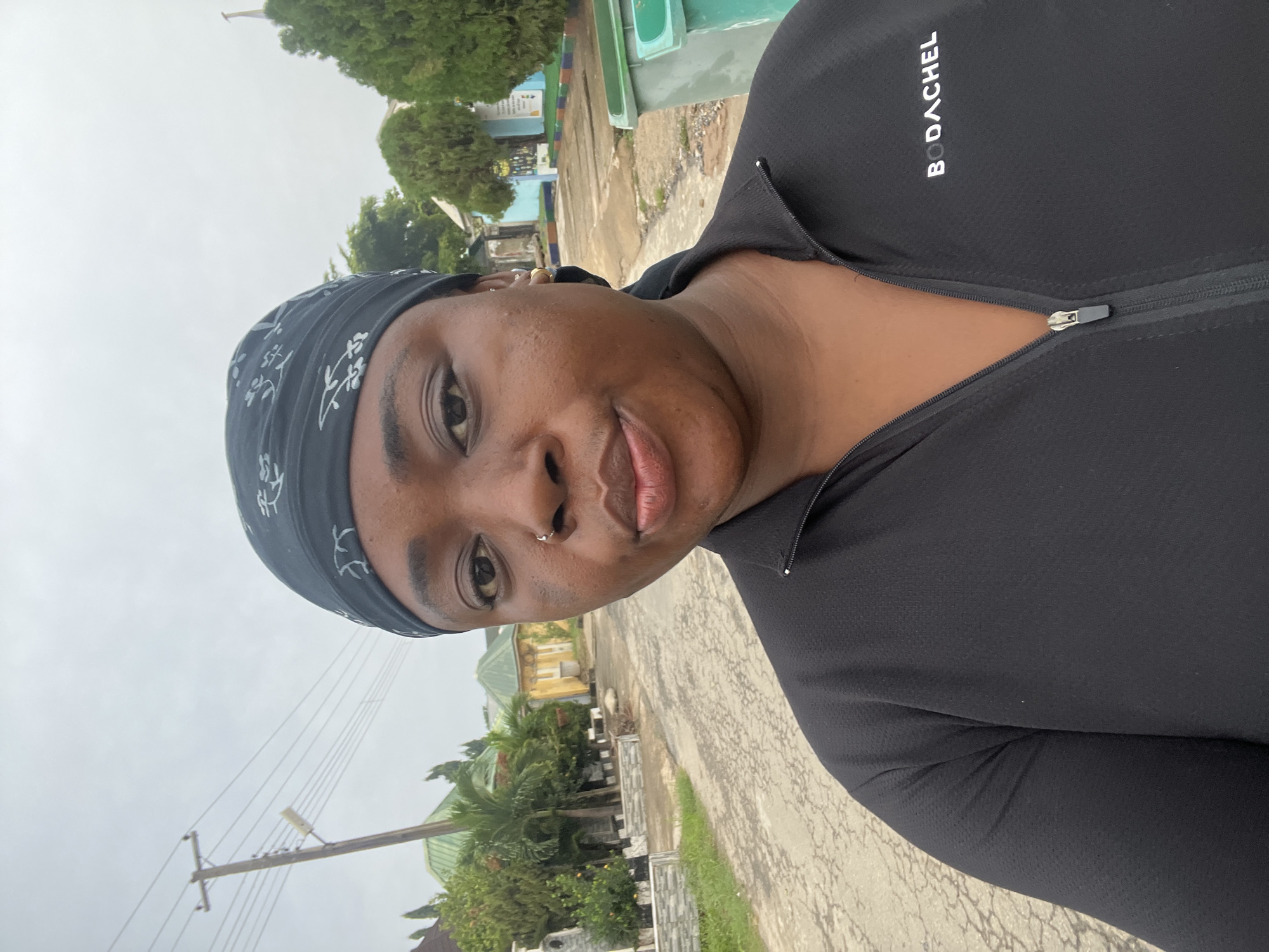 A person wearing a black headband and a black athletic jacket with a partial zipper is the primary subject of the image. The jacket has the word "BODACHEL" printed on it. The person has a nose ring and is likely taking a selfie.
The background shows a street scene in Abuja, Nigeria, with residential buildings and green vegetation lining the sides of the road. There is also a utility pole with overhead wires visible in the distance. The sky is overcast. The pavement appears to be concrete or asphalt.