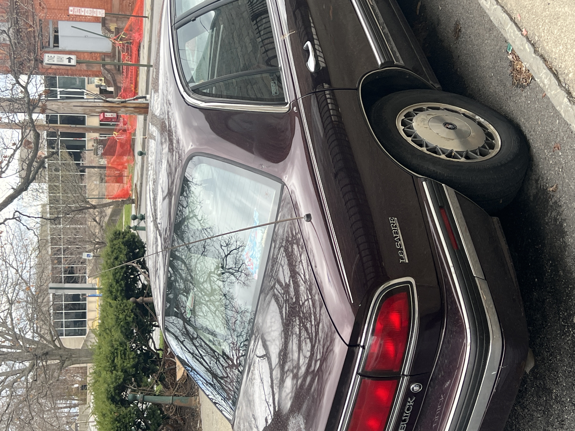 The image shows a maroon Buick LeSabre parked on a street in the City of Newburgh, United States of America. A building undergoing construction and a "One Way" street sign are visible in the background. The reflection of bare trees is visible on the car's rear window.