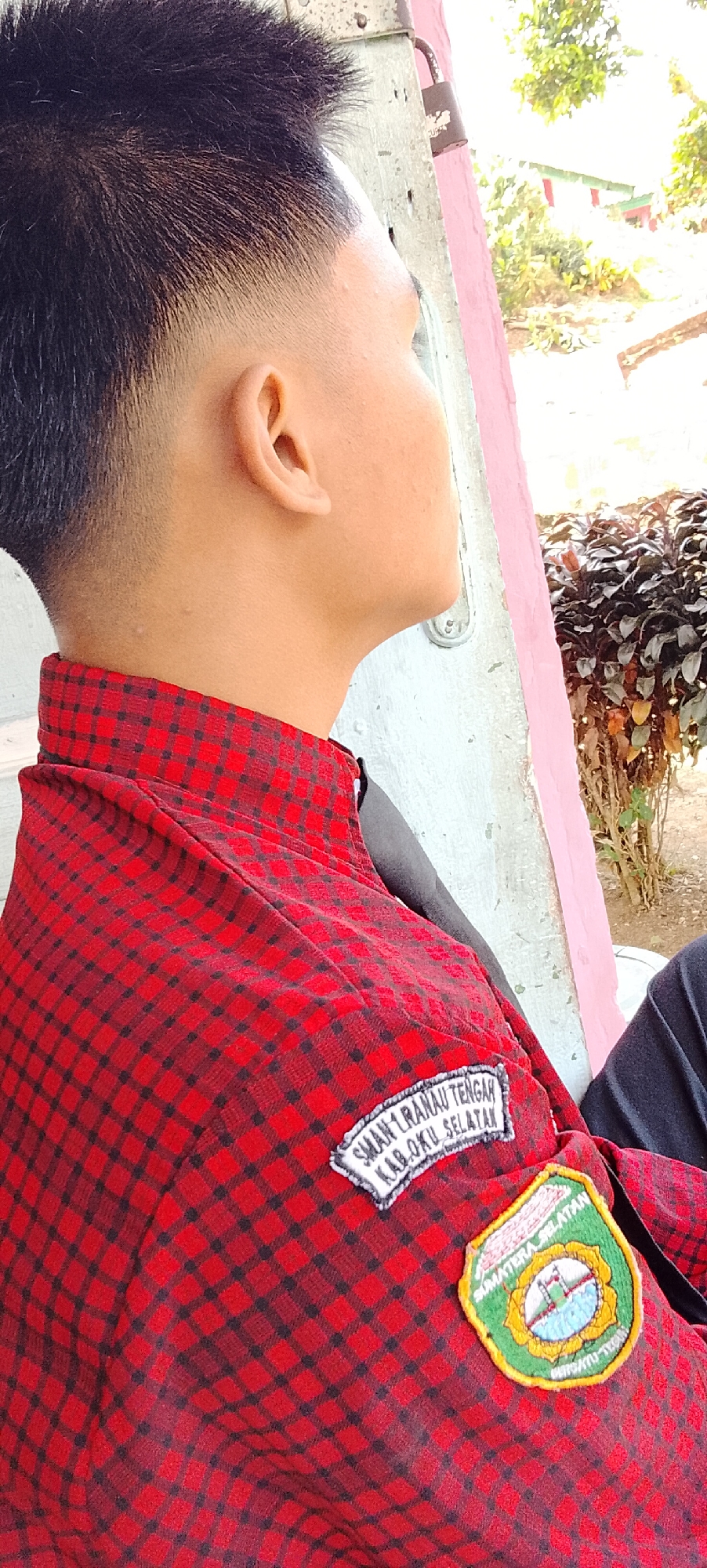 A young male is shown in profile, looking to the right. He has a fresh haircut with a fade on the sides. He is wearing a red and black checkered shirt with two patches on the left side of his chest. The top patch is white with black lettering that reads "SMAN 1 RANAU TENGAH KAB. OKU SELATAN". Below this is a circular patch with a green border and a design in the center that appears to be a landscape with water and trees. The background is out of focus, but suggests an outdoor setting with foliage and what looks like a building with a pink wall. The image is taken from a medium close-up, focusing on the upper torso and head of the subject. The lighting suggests it is daytime, possibly sunny. Given the text on the patch, it is highly probable this image was taken in or near Tanjung Setia, Indonesia, as this refers to a specific school in that region.