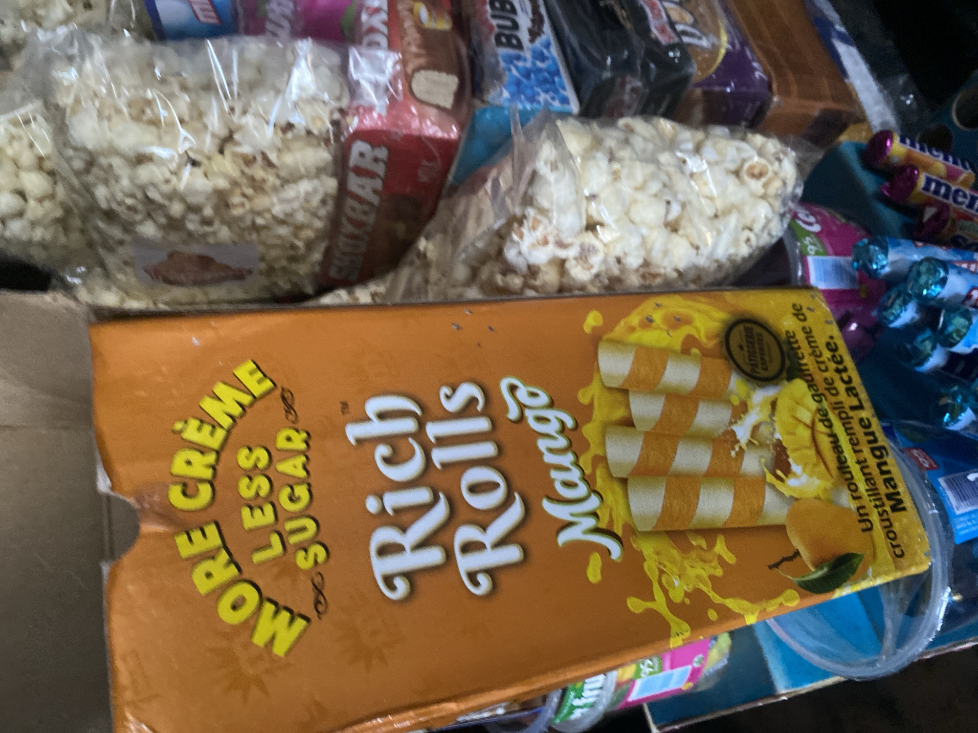 The image shows a close-up of a stack of snacks, primarily popcorn and chocolate bars, with a box of "More Creme Less Sugar Rich Rolls Mango" in the foreground. The snacks are packaged in clear plastic and colorful wrappers. The setting appears to be a retail environment, possibly a market or a store shelf. The lighting is artificial and casts shadows, suggesting an indoor location. There are no people or any specific environmental features visible that would indicate a precise location within Lafia, Nigeria.
