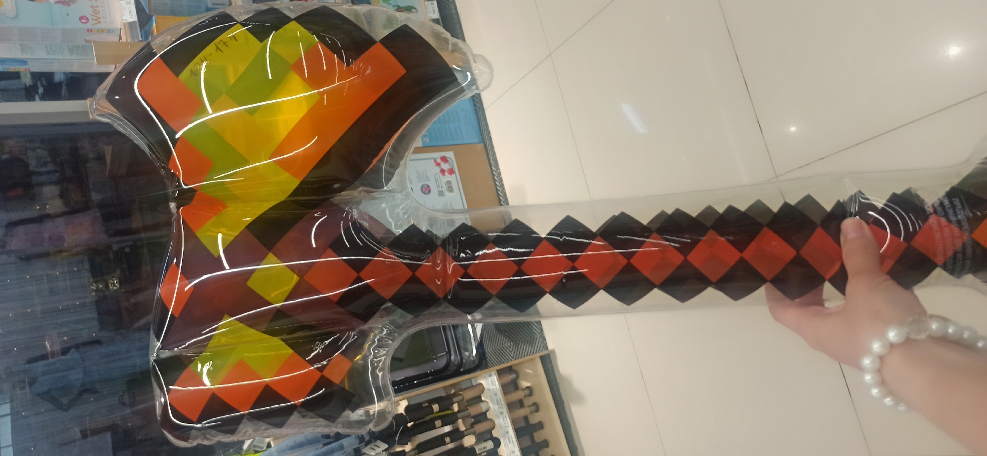 In the city of Asbest, Russia, an individual's hand, adorned with a pearl bracelet, is holding a large, inflatable toy axe. The axe has a diamond pattern with alternating black, orange, and yellow sections on its head, and a black and orange diamond pattern on its handle. The background suggests an indoor retail environment, with shelves stocked with various items, including what appear to be tools or similar merchandise, and a display of clothing or mannequins behind a glass partition. The floor is tiled and appears to be a light color.