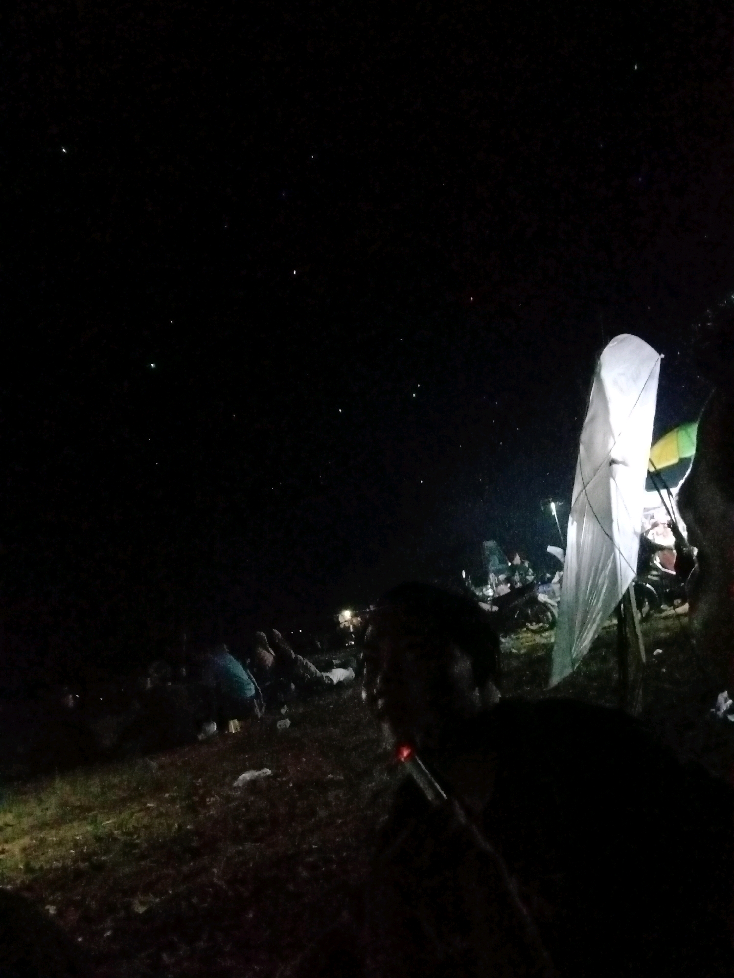 The image depicts a nighttime outdoor gathering, likely in Qurnia Mataram, Indonesia, given the context provided. The scene is dark, with a few scattered lights visible in the sky, possibly stars or distant lights. In the foreground, a person is silhouetted against the dim light, appearing to be smoking a cigarette.
In the background, a large, white, lantern-like object is prominent, possibly a sky lantern or a decorative light. Several other people are visible, some seated or lying down, suggesting a relaxed or resting atmosphere. A few motorcycles are parked nearby, and there appears to be some debris scattered on the ground. The environment seems to be an open area, possibly a field or a cleared space, with some vegetation visible. The overall impression is a casual nighttime event or gathering.