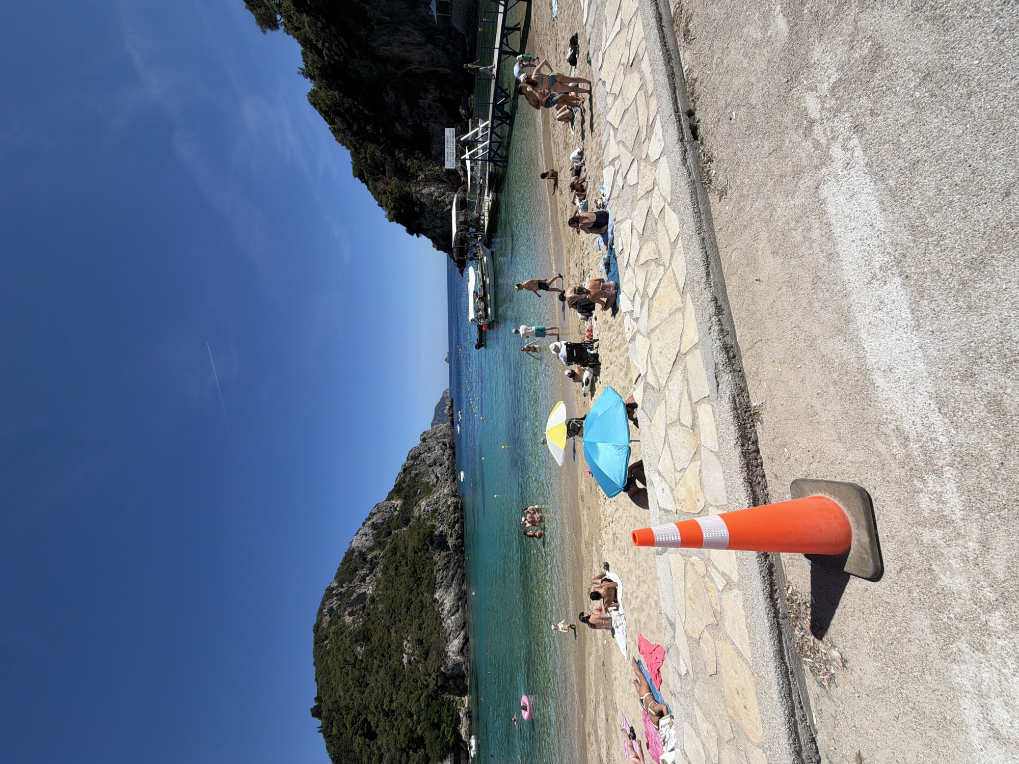 This detailed image depicts a bustling beach scene in Palaiokastritsa, Greece, under a clear, bright blue sky with a faint contrail visible in the upper right. The bay is framed by steep, verdant cliffs with exposed rock faces. The water is a clear, calm turquoise, with yellow buoys marking off a swimming area.
The sandy beach is populated by approximately 25-30 individuals engaged in various recreational activities. On the left side of the beach, several people are sunbathing on colorful towels (pink, white) directly on the sand. A child in a pink inflatable ring is floating in the shallow water nearby. Towards the center, multiple groups of people are standing or sitting. Two beach umbrellas are prominent: one yellow and white, and another solid blue. A wheelchair and a baby carriage are visible on the left side of the beach.
In the water, roughly 10-15 individuals are swimming, standing, or interacting in groups. On the right side of the bay, a wooden pier with a covered section extends into the water. Two white boats are docked at this pier, and several people are seen on and around the structure. A sign with text in Greek is affixed to the pier's shelter.
In the immediate foreground, a textured, light-colored stone-paved walkway transitions to a gray concrete road. An orange traffic cone with two white reflective stripes stands prominently on the concrete road in the lower-center, casting a distinct shadow to its left. The concrete road shows signs of wear, and a faint white line is visible on its surface to the far right. Debris is visible at the junction of the stone paving and the concrete road. The entire scene is brightly illuminated by direct sunlight, creating sharp shadows.