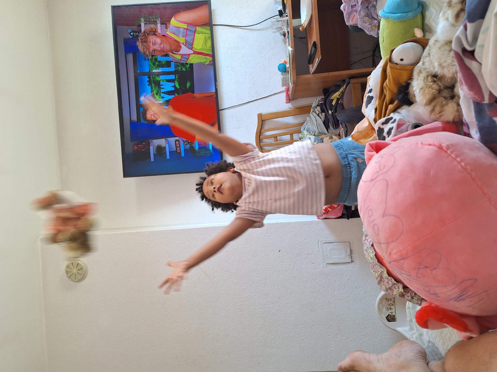 The image depicts an indoor scene, possibly a bedroom, where a young child with curly hair is in the process of throwing a stuffed toy into the air. The child is wearing a striped t-shirt and denim shorts. In the background, a television screen displays two men, one in a red shirt and the other in a neon vest. A wooden chair and a cluttered desk with various items are also visible. The environment is brightly lit. There are no identifiable elements that would suggest the location is Blagoevgrad, Bulgaria.