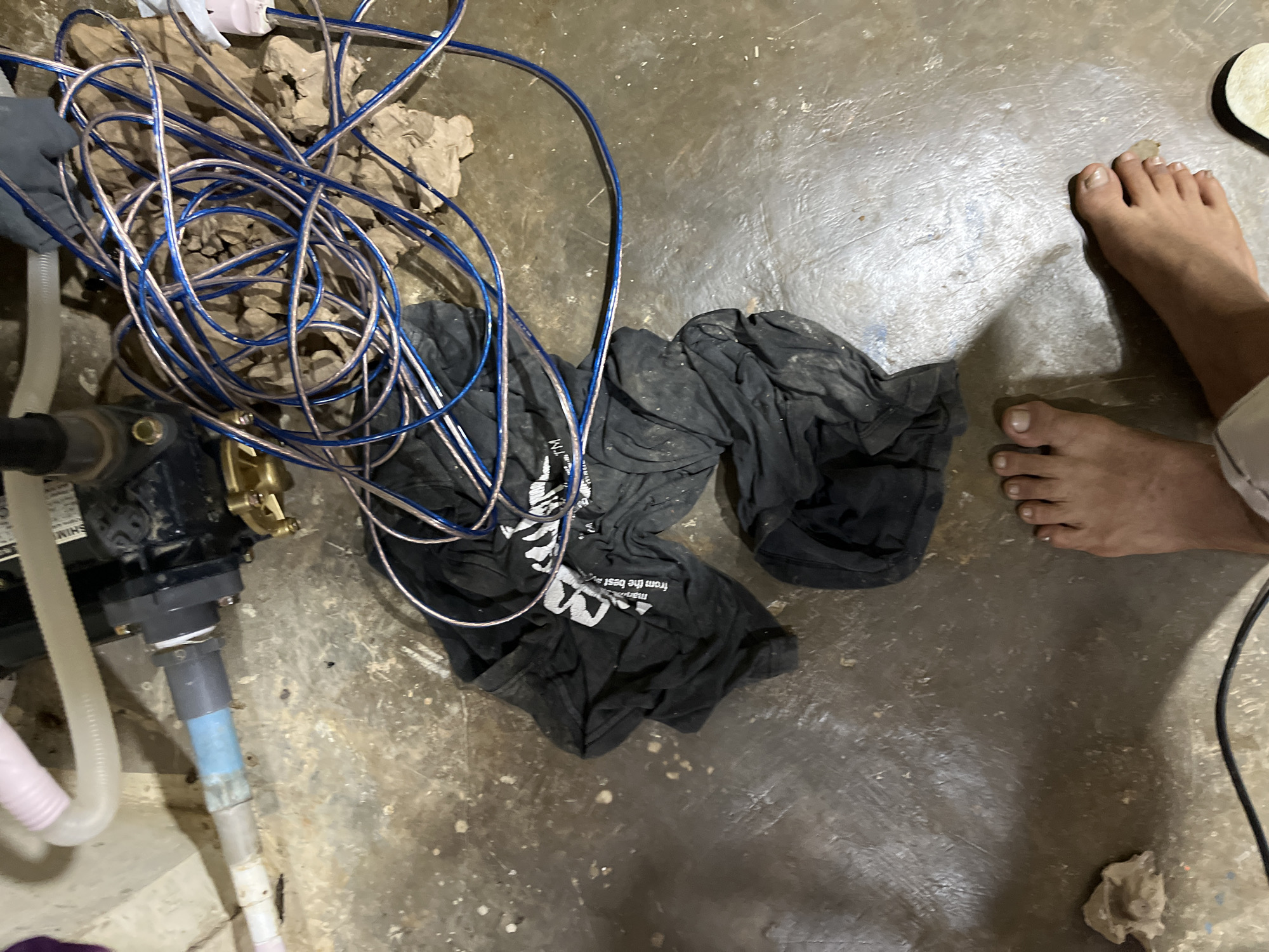 A close-up, overhead shot shows a man's bare feet on a damp, concrete floor in what appears to be a basement or utility area. The floor is marked with white splatters and some dirt. To the left of the feet, a pile of dark fabric, possibly a t-shirt, is crumpled on the ground, with some visible white lettering that is partially obscured. A tangled mess of blue and copper wires is draped over the fabric and extends towards a dark-colored pump or motor with brass fittings. A light-colored, corrugated pipe is visible in the upper left corner, connected to a grey pipe with blue tape. The image is devoid of any elements that would indicate a specific location within the city of Benuaraja, Indonesia.