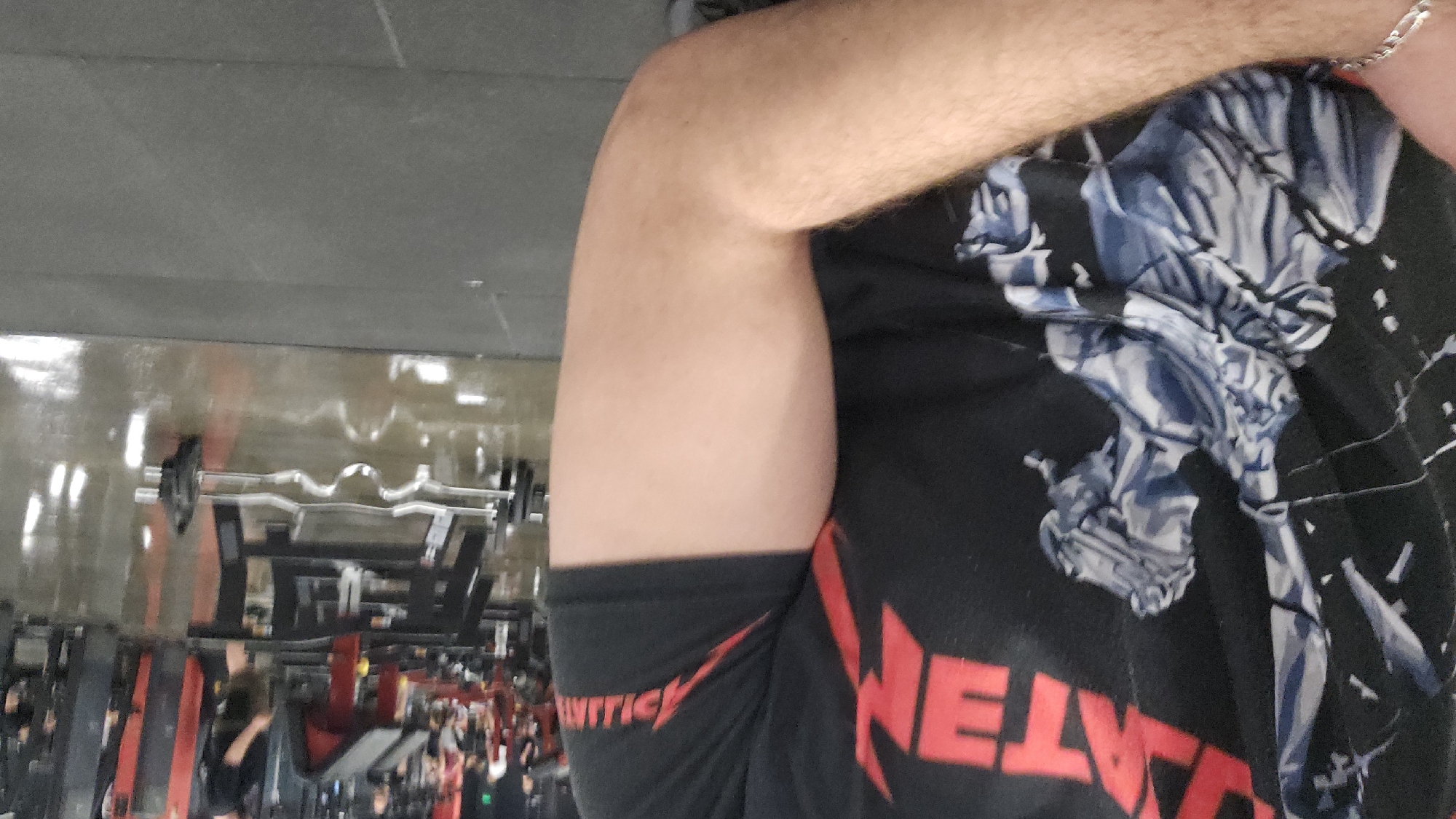 The image, rotated 90 degrees clockwise, captures a partial view of a person's upper body and arm within a gym setting in Plovdiv, Bulgaria. The subject, likely male, is prominently displaying their right arm, suggesting a flexed bicep, indicating physical activity or a pose. A silver, braided bracelet is visible on their wrist.
They are wearing a black short-sleeved t-shirt with red and blue-grey graphics. Visible text on the t-shirt includes "VNTTICK" with a lightning bolt design on the left sleeve or side, and a large red "METALLICA" logo prominently displayed on the torso. A detailed blue-grey graphic of a skeletal figure is also present on the front of the shirt. The skin on the arm is light-toned with visible hair.
The background, out of focus due to the close-up shot, reveals the interior of a fitness center. The floor, appearing on the left side of the rotated image, is glossy and reflective, showing streaks and lights, along with some red markings. Various gym equipment is discernible, including silver barbells (one appears to be an EZ curl bar), weight racks, and exercise benches. Blurry figures of other individuals are visible in the deeper background, engaged in activities around the equipment. The ceiling, or what appears to be the ceiling on the right side of the rotated image, is a plain, light grey concrete-like surface.