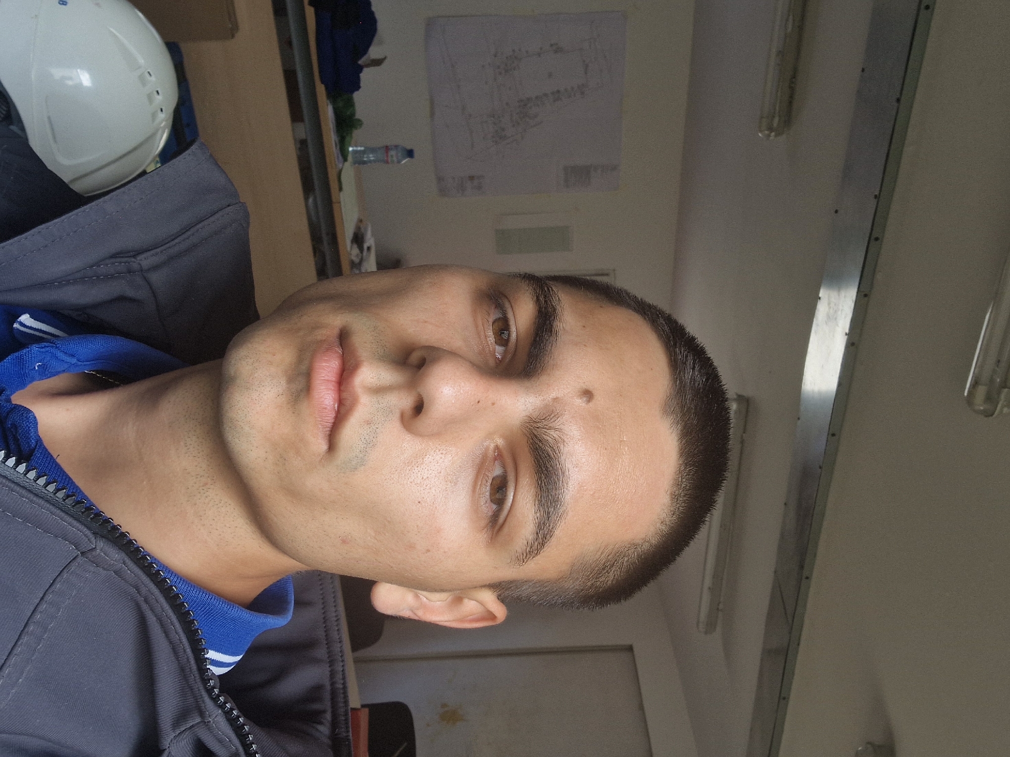 A selfie photograph features a person with a short, dark haircut, tan skin, and brown eyes. They are wearing a blue collared shirt under a dark gray jacket with a zipper. A white hard hat is visible to the upper left of the frame. In the background, there is a desk with various items, including a water bottle, a paper with technical drawings, and some clothing. The background is a pale, possibly industrial or office space, with fluorescent lights and a plain wall. The photograph is slightly rotated, so the scene appears tilted. There is no clear indication of the exact location in Sofia, Bulgaria.