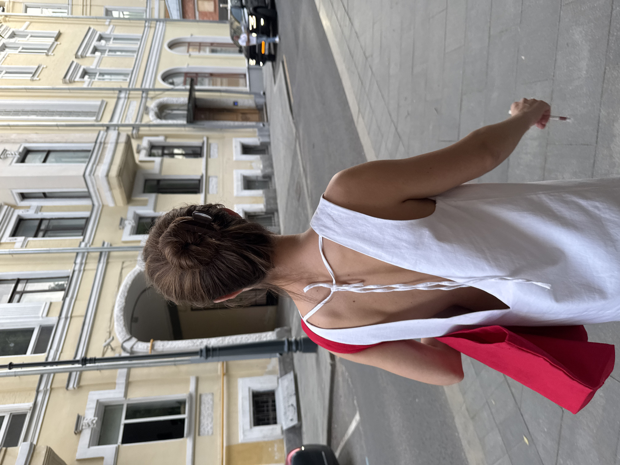 A medium-shot, angled, eye-level photograph shows a woman with a dark bun walking down a city street. The woman is wearing a white dress with tie straps in the back and has a red bag hanging off one shoulder. She has a slender arm that is holding what looks like a cigarette, and is facing towards the camera, although the back of her head is mostly visible. The image is taken on a grey sidewalk next to a street with cars visible. On the other side of the street is a tall, yellow building with white trim and many square windows. The photograph was taken in Moscow, Russia.