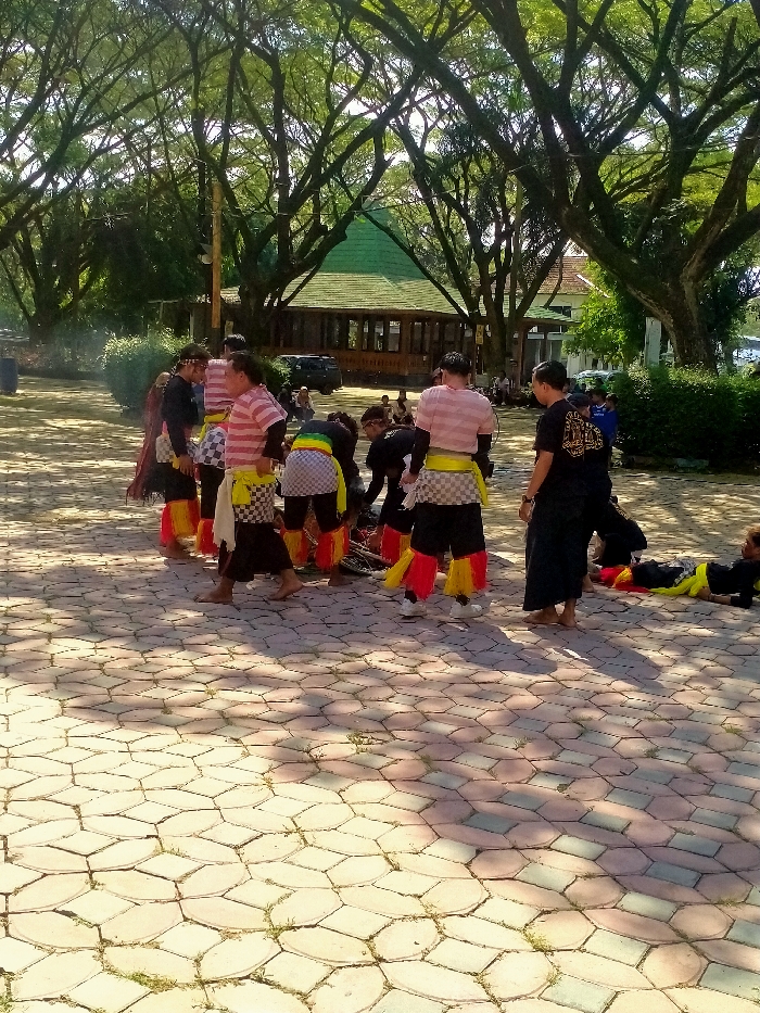 A group of people in traditional attire are gathered in an outdoor setting. Some wear red and yellow leg decorations, striped shirts, and checkered belts. They are positioned on a paved ground with an octagonal pattern. Lush green trees provide shade, and a building with a distinctive green roof is visible in the background. Several other people are watching the gathering. The scene is captured under daylight conditions.