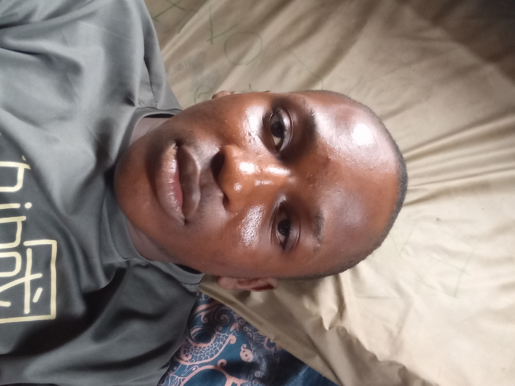 A close-up, over-the-shoulder shot captures the face of a young Black person lying on a surface, possibly a bed, in Nkpor, Nigeria. The person is wearing a grey t-shirt with a light-colored graphic design on the left side of the chest. Their face is turned towards the camera, with their head tilted upwards and to the right. Their skin appears to be oily or sweaty, reflecting the light. Their lips are slightly parted, and their eyes are open. The background consists of a tan-colored sheet or blanket, with some faint lines or creases visible. To the lower right of the image, a patterned fabric, possibly part of a bedspread, is visible. The overall lighting suggests it may be indoor lighting.