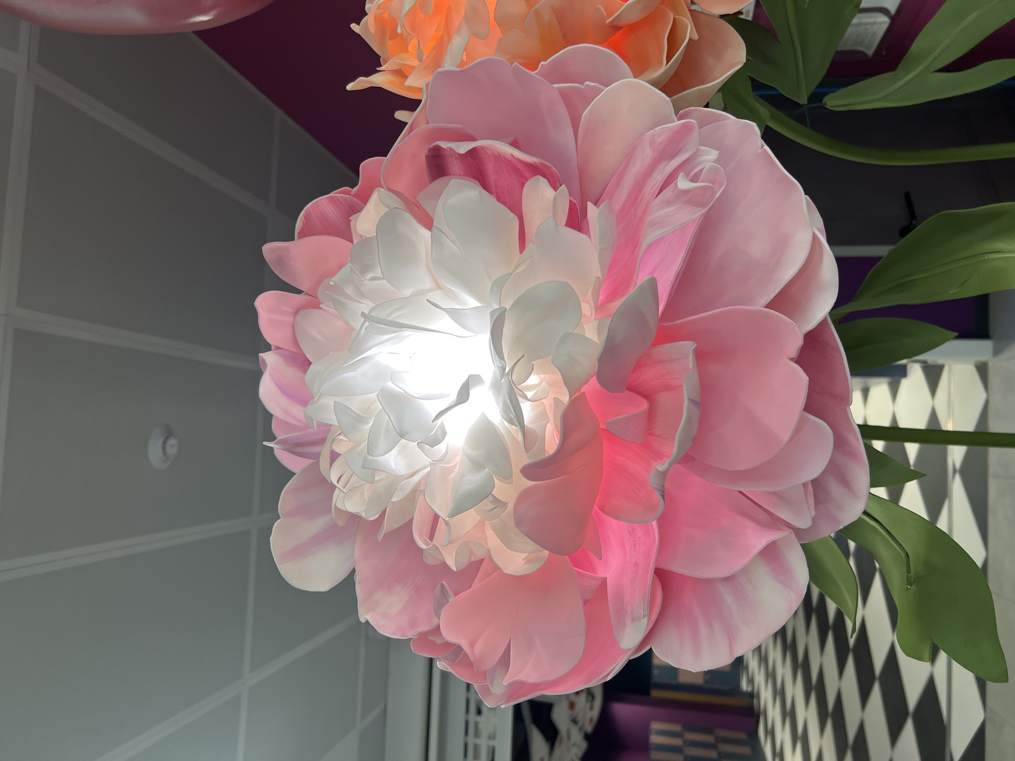 This is a close-up, low-angle shot of a large, illuminated, artificial pink and white flower. The flower's petals are layered and translucent, with a bright light source emanating from the center. To the right, a portion of another large artificial flower, colored orange, is visible. The background is out of focus and features a black and white checkered floor, a purple wall, and a grey ceiling with white grid lines. There are no people or animals in the image. The location is Vaganovo Massiv, Russia.