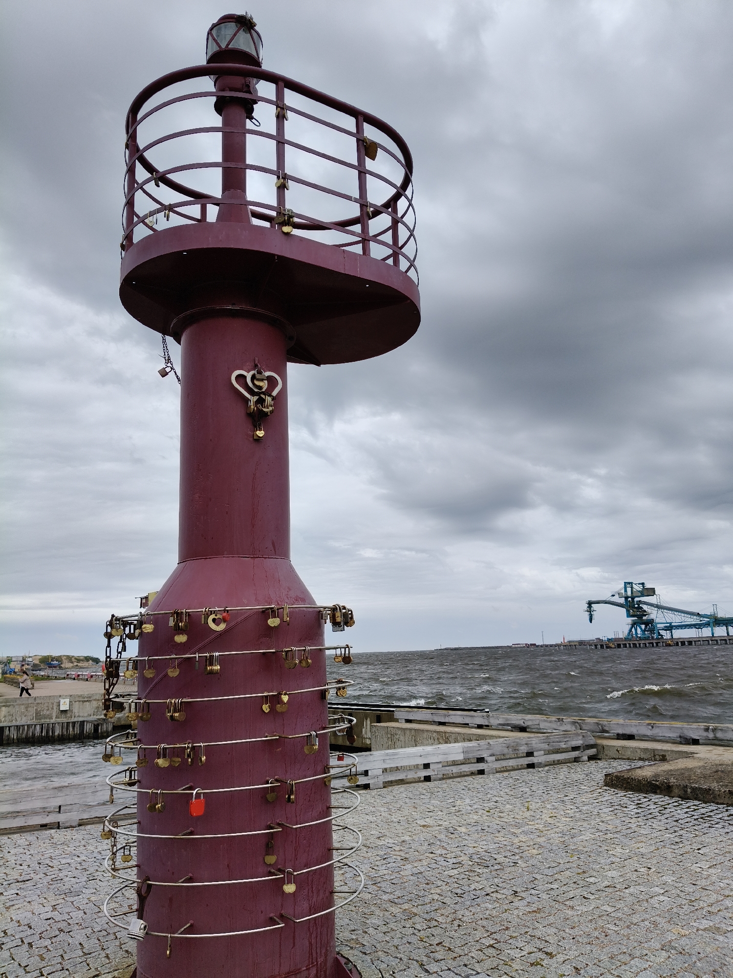 A lighthouse with a red casing stands prominently in the foreground, adorned with numerous padlocks attached to its railings and structure. The lighthouse has a circular viewing platform near its top, topped by a lantern room with a glass enclosure. Below the platform, the lighthouse tapers downwards and is wrapped in horizontal metal bars where more padlocks are secured. One bright red padlock stands out among the many brass-colored ones.
To the left of the lighthouse, a paved walkway leads towards a wooden pier. A solitary person is visible walking on this pier. The sea, with choppy waves, stretches into the background, meeting a cloudy, overcast sky. On the far side of the sea, industrial structures, including a large blue crane, are situated along the Ventspils coastline. The ground in the immediate foreground is paved with gray cobblestones. The scene appears to be a public waterfront area in Ventspils, Latvia, likely a harbor or a pier.