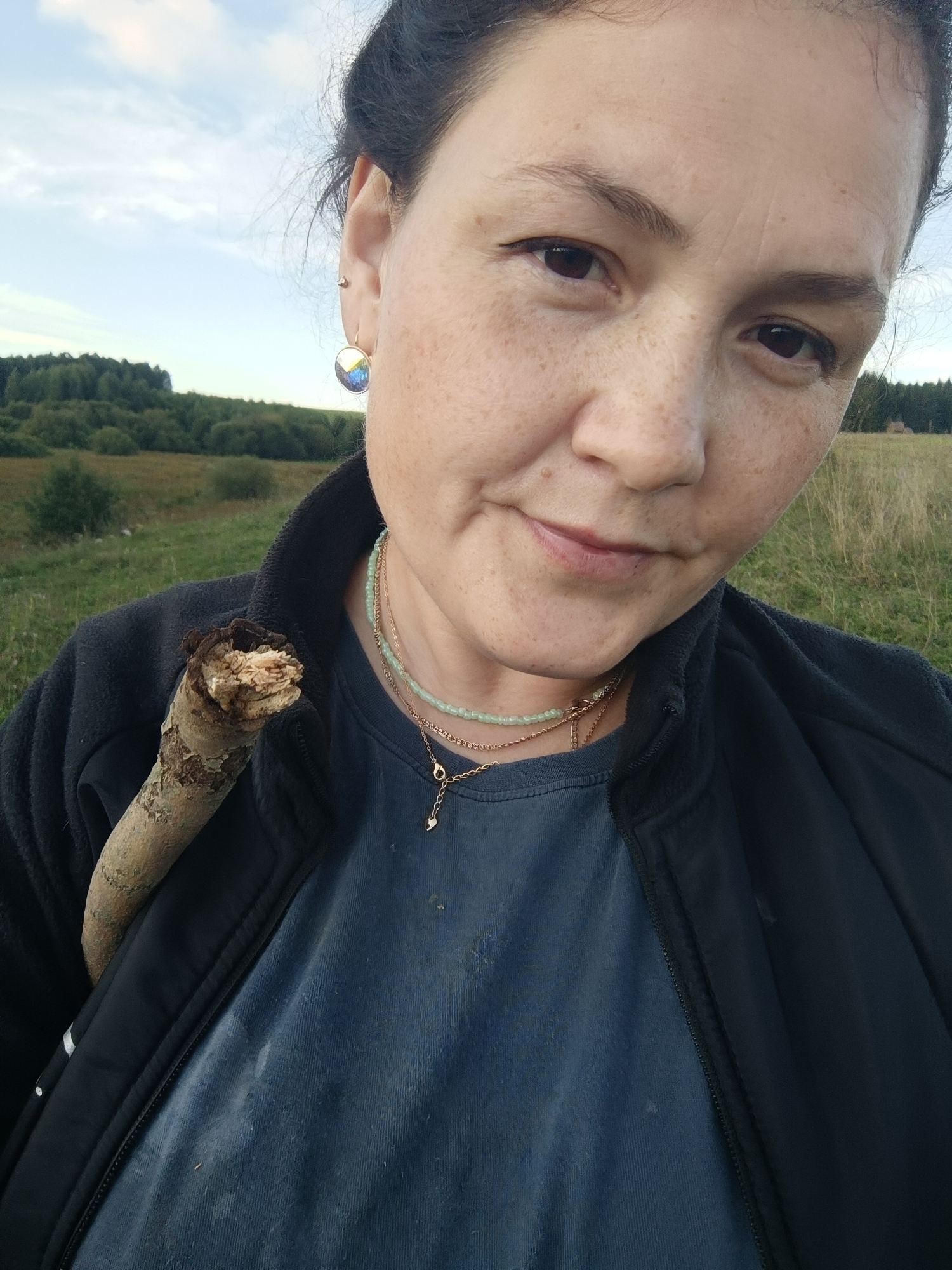 In a frontal, medium close-up shot, a woman with dark hair pulled back and visible freckles across her face is pictured outdoors. She is wearing a dark blue collared jacket over a dark blue t-shirt, adorned with two necklaces, one a strand of light green beads and the other a delicate gold chain with a small charm. A broken wooden stick is held diagonally across her left shoulder and chest. The background shows a slightly blurred landscape with a treeline and a cloudy sky, suggesting a rural or natural setting. No specific location in Karsha, Russia is discernible from the image.