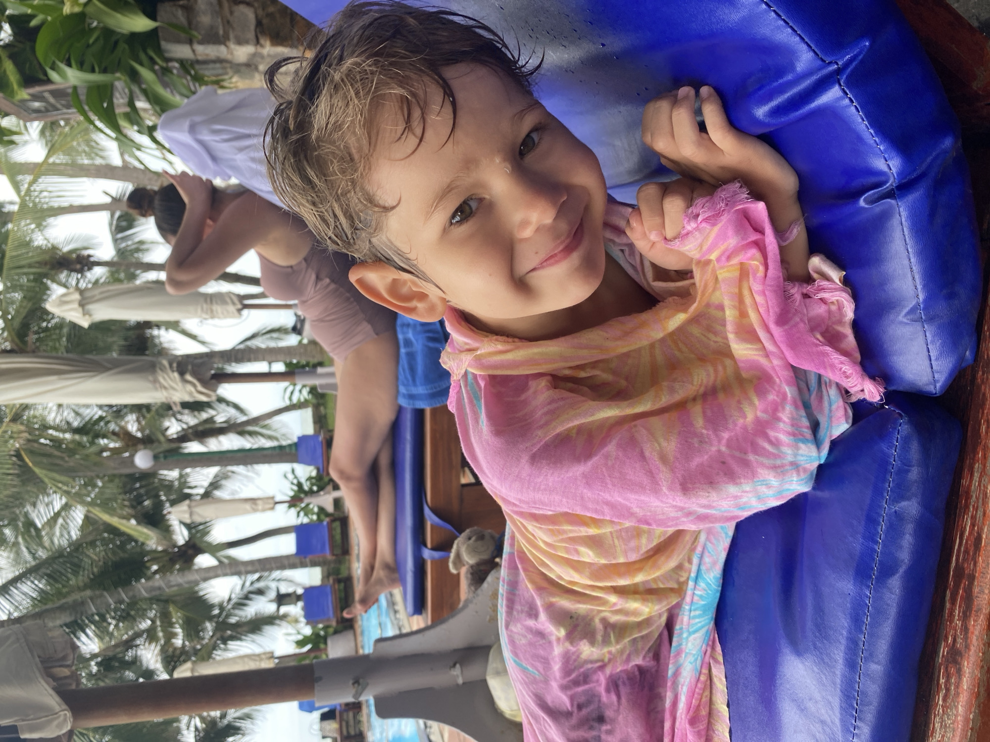 A young child with wet, light brown hair and green eyes smiles at the camera, wrapped in a pink and yellow tie-dye towel. The child is lying on a bright blue padded lounge chair. In the background, another person's legs are visible, stretched out on a similar chair, with a swimming pool and palm trees suggesting a resort or hotel setting in Phu Thuy Ward, Vietnam. The scene appears to be outdoors, possibly near a beach or pool area, with a slightly overcast sky.