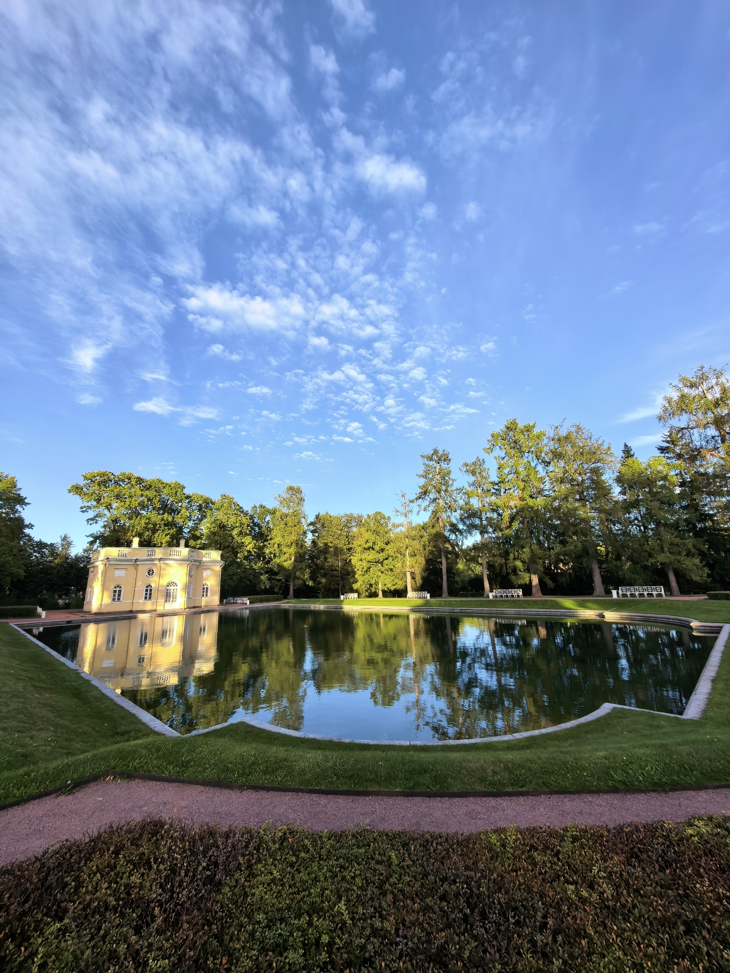 The image depicts a scenic view of the Catherine Park in Pushkin, Russia, featuring a tranquil pond reflecting a bright yellow building and the surrounding trees. The sky above is a vivid blue, adorned with scattered, wispy white clouds. The building, with its classical architecture and numerous windows, is situated to the left of the frame, its reflection clearly visible in the calm water. A lush green lawn borders the pond, with a gravel path and manicured bushes in the foreground. Tall, mature trees line the far side of the pond, their green foliage creating a vibrant backdrop. Several white benches are visible along the treeline, suggesting a place for public relaxation. The overall impression is one of serene natural beauty and historical architecture.