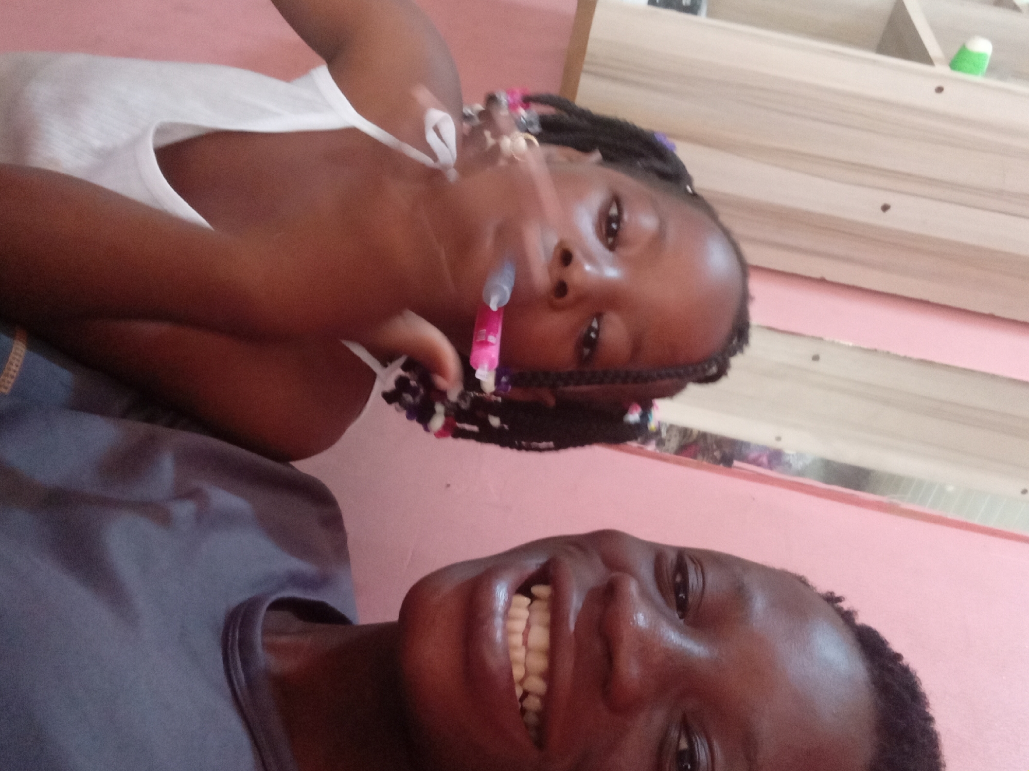 Two young Black individuals are in a close-up selfie. The person on the right, closer to the camera, is smiling broadly, showing their teeth. Their head and shoulders are visible. The person on the left, slightly further back, is looking towards the camera with their mouth open, holding a pink and grey object, possibly a toy syringe, near their mouth. This individual has braided hair with colorful beads. Both are wearing casual clothing. The background appears to be an indoor setting with a pink wall and wooden paneling. The lighting suggests an indoor, possibly domestic, environment.