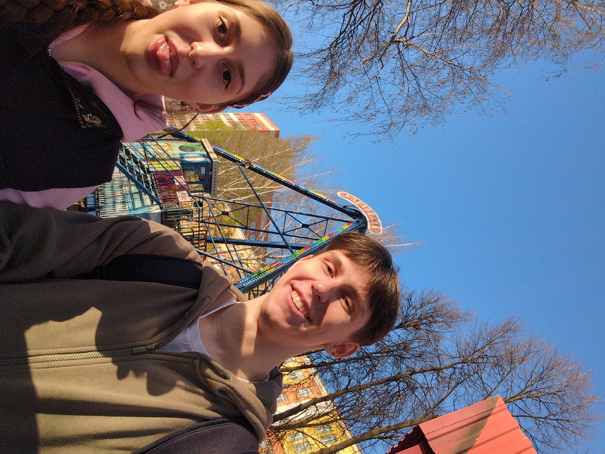 Two individuals, a woman and a man, are positioned in the lower left and right corners of the frame, respectively, in an outdoor environment. The woman, at the top left, has brown hair, fair skin, and is sticking her tongue out. She is wearing a pink shirt under a black jacket. The man, at the bottom right, also has brown hair and fair skin, and is smiling. He is wearing a white t-shirt underneath a green zip-up jacket and has a backpack slung over his shoulder.
In the background, partially obscured by the subjects, is a Ferris wheel with colorful decorations, and buildings with light yellow and orange facades with windows. Bare branches from trees are visible against a clear, bright blue sky. Based on the Ferris wheel design and building architecture, the location is Izhevsk, Russia.