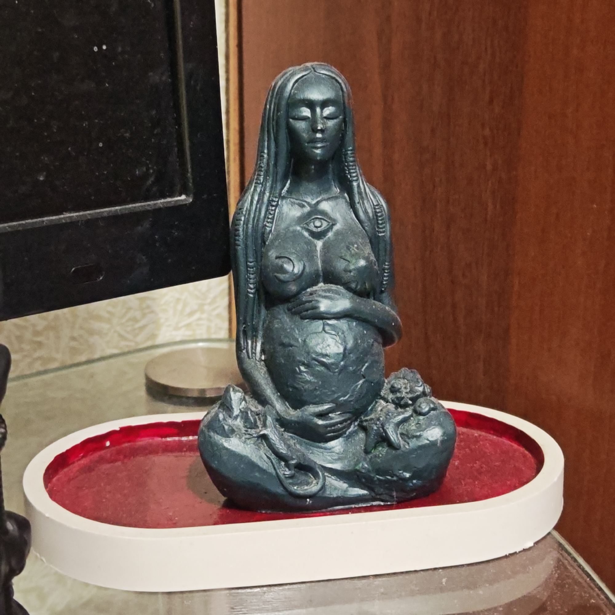 The image displays a dark, possibly charcoal grey or dark green, statue of a pregnant woman, situated in an indoor setting in Ketoshi, Russia. The statue depicts a figure with long, textured hair cascading over her shoulders and closed eyes, suggesting a serene or meditative state. A prominent "third eye" symbol is carved on her chest, along with a crescent moon symbol below her left breast. Her hands are gently placed over her visibly pregnant belly. The woman is seated in a cross-legged or lotus-like pose, with intricate, organic textures and shapes around her base.
The statue rests on a distinctive white, oval-shaped tray with a vibrant red, slightly textured or dusty recessed interior. This tray is positioned on a glass table. To the immediate left, a portion of a dusty black electronic device, resembling a monitor or television screen, is visible. The background features warm-toned wooden paneling or furniture and a light-colored, subtly patterned wallpaper. The lighting suggests an indoor, possibly domestic environment. No discernible activity is taking place beyond the static display of the artwork. No text is visible in the image. The overall impression is one of calm, spiritual maternity.
