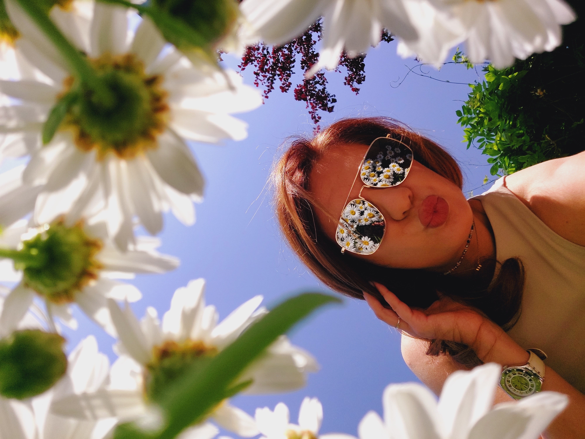 A young woman with auburn hair is photographed from a low angle, surrounded by white daisies against a clear blue sky. She is wearing sunglasses that reflect a field of daisies, and her lips are pursed in a kiss. Her right hand is touching her hair, and she is wearing a ring on her index finger. She also has a necklace and a watch with a green face on her left wrist. In the background, there is a tree with dark red leaves and green foliage. The image appears to be taken outdoors, possibly in a garden or park in Sofia, Bulgaria, given the bright sunlight and the presence of flowers and greenery.