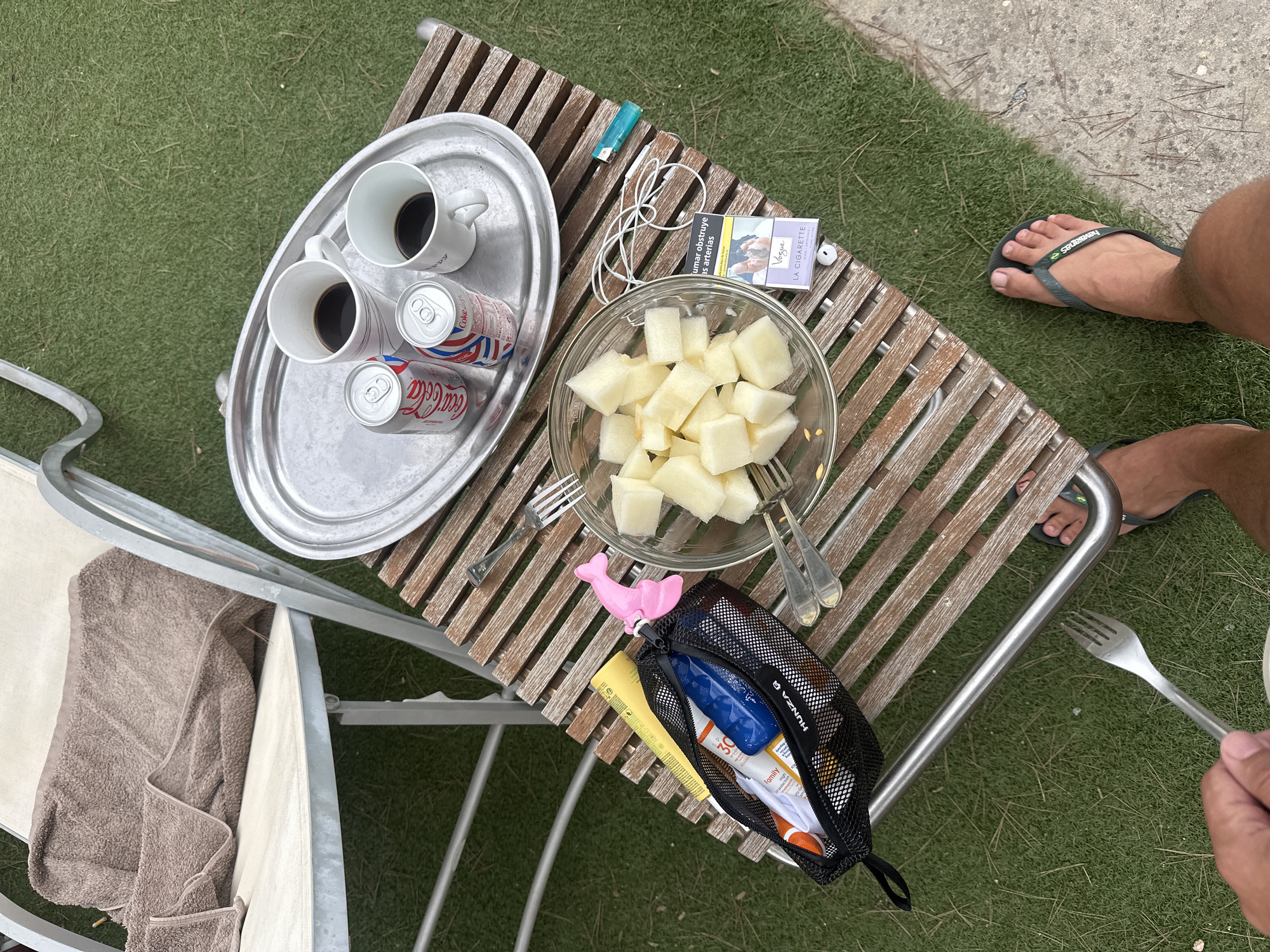 An overhead shot shows a brown towel draped over a portion of a light-colored lounge chair with a metal frame. The chair is situated on a patch of artificial grass. To the right of the chair, a wooden table with horizontal slats holds a silver tray. On the tray are two white mugs with dark liquid, two cans of Coca-Cola, and one can of Coke Light. A clear glass bowl filled with cubed melon sits on the table, accompanied by two forks. A black mesh bag, containing items such as a yellow ruler, a blue bottle, a tube of sunscreen, and a white shirt, is also on the table. A pink plastic dolphin-shaped object is attached to the zipper of the bag. Further to the right on the table, a pack of Vogue cigarettes and a teal lighter are visible. A white cord with earbuds is also present. Below the table, a person's feet are visible wearing dark green flip-flops. One person is holding a fork and is partially visible in the lower left corner.
