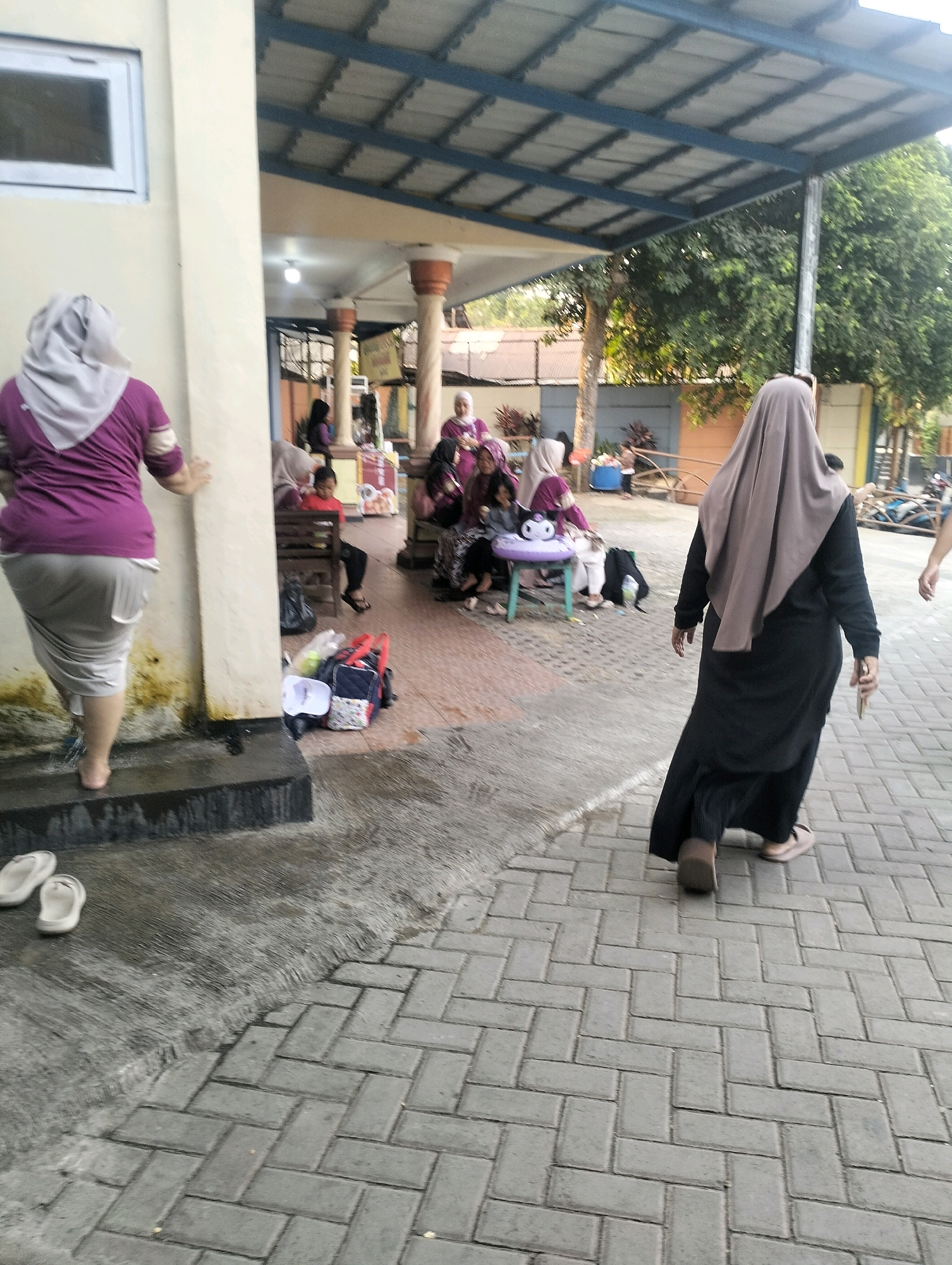 A group of people, primarily women wearing hijabs, are gathered in an outdoor covered area. The area appears to be part of a public facility, possibly a school or community center, with a tiled floor and a corrugated metal roof supported by pillars. In the foreground, a woman in a purple top and a light-colored skirt is standing near a concrete wall, her hand resting on it. To her right, a woman in a dark long dress and a beige hijab walks away from the camera. In the background, several women are seated on benches, some with children. Bags and other items are scattered on the ground. The environment suggests a warm, sunny day. The overall scene depicts a casual gathering.
