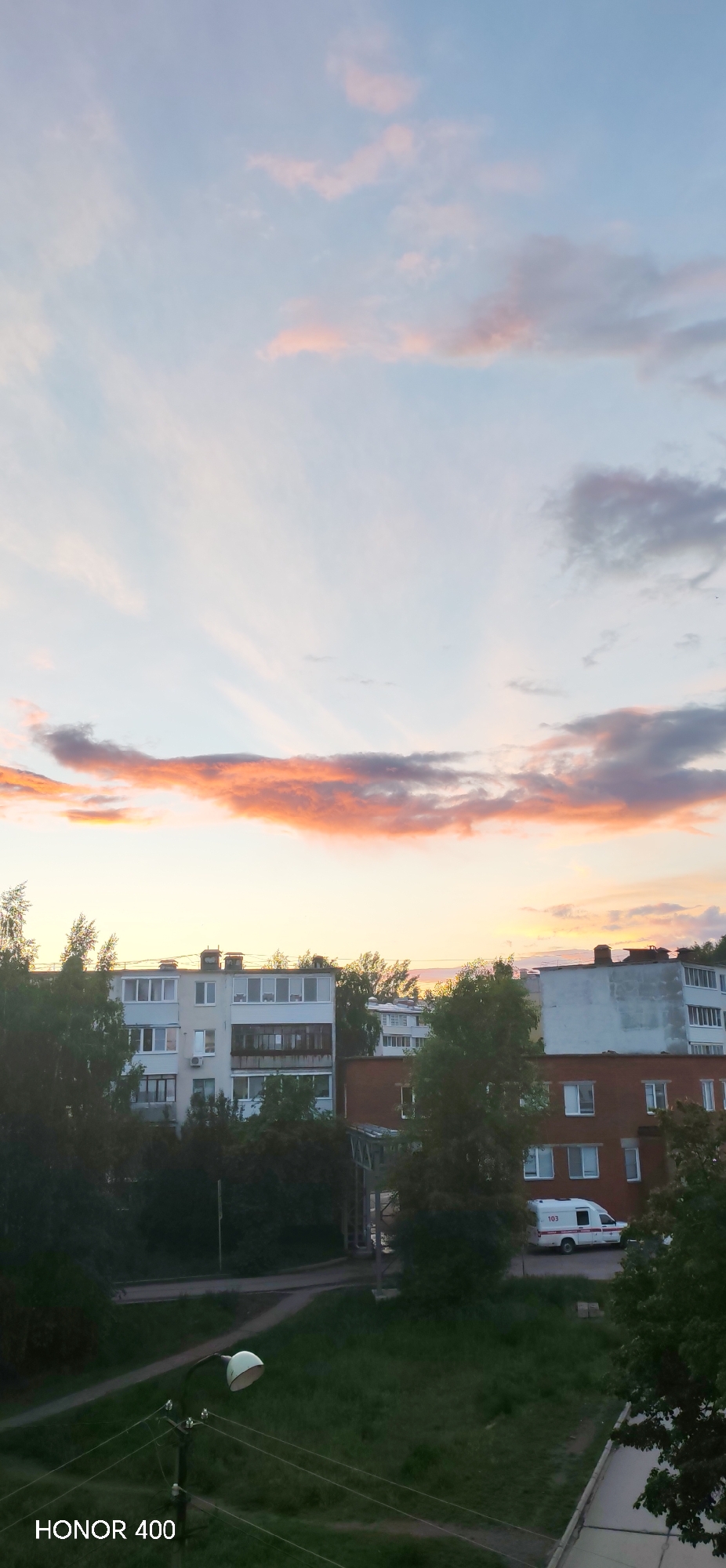 The image depicts a cityscape under a colorful sunset sky. Predominantly, the sky exhibits hues of pink, orange, and grey clouds spread across a pale blue background. Below the horizon, several buildings are visible, varying in color and architecture. The trees partly obscure the buildings, filling the gaps with green foliage. In the lower part of the picture, there is a green space. Near the buildings, an ambulance is parked, marked with '103'. The image was taken in Noviy, Russia. The bottom left corner contains the text "HONOR 400".
