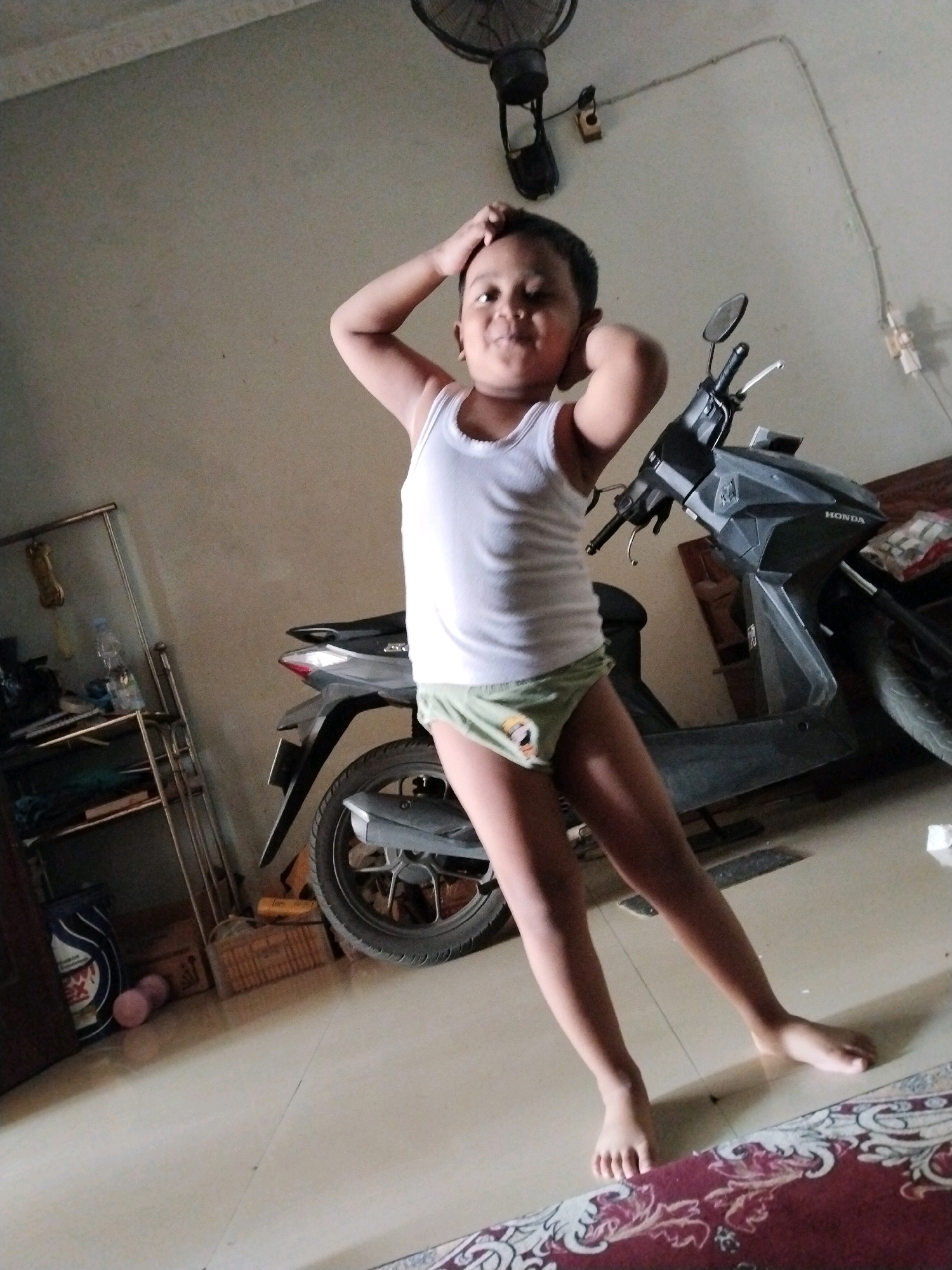 A young boy is standing in what appears to be a room, possibly a garage or a covered outdoor area, in Pepelegi, Indonesia. He is wearing a white tank top and light green shorts with a cartoon character on them. The boy has his hands on his head, striking a playful pose.
Behind him, a dark-colored scooter is parked. The scooter has a "HONDA" logo visible on its side. To the left of the scooter, there is a metal shelving unit with various items on it, including a plastic bottle of water. The floor is tiled and a portion of a rug with a patterned design is visible in the foreground. A ceiling fan is mounted on the wall above the boy. The lighting suggests it could be daytime. The overall impression is one of a casual moment captured at home.