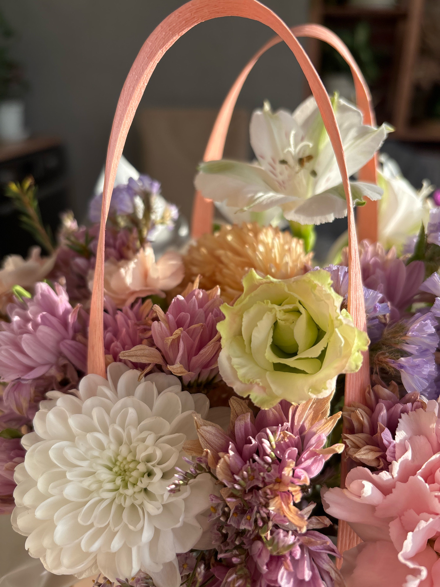 This is a close-up, high-angle, natural light photograph of a bouquet of flowers in a basket with pink handles. The flowers are varied in color and type, including white chrysanthemums, pale yellow roses, pink carnations, purple statice, and some smaller, delicate white flowers with green centers. The background is blurred, showing hints of a dark wall and wooden shelving. The scene is an indoor still life, likely a domestic setting. There are no people or animals present. The photograph does not provide any specific information about the location in the city of Sapronovo, Russia.