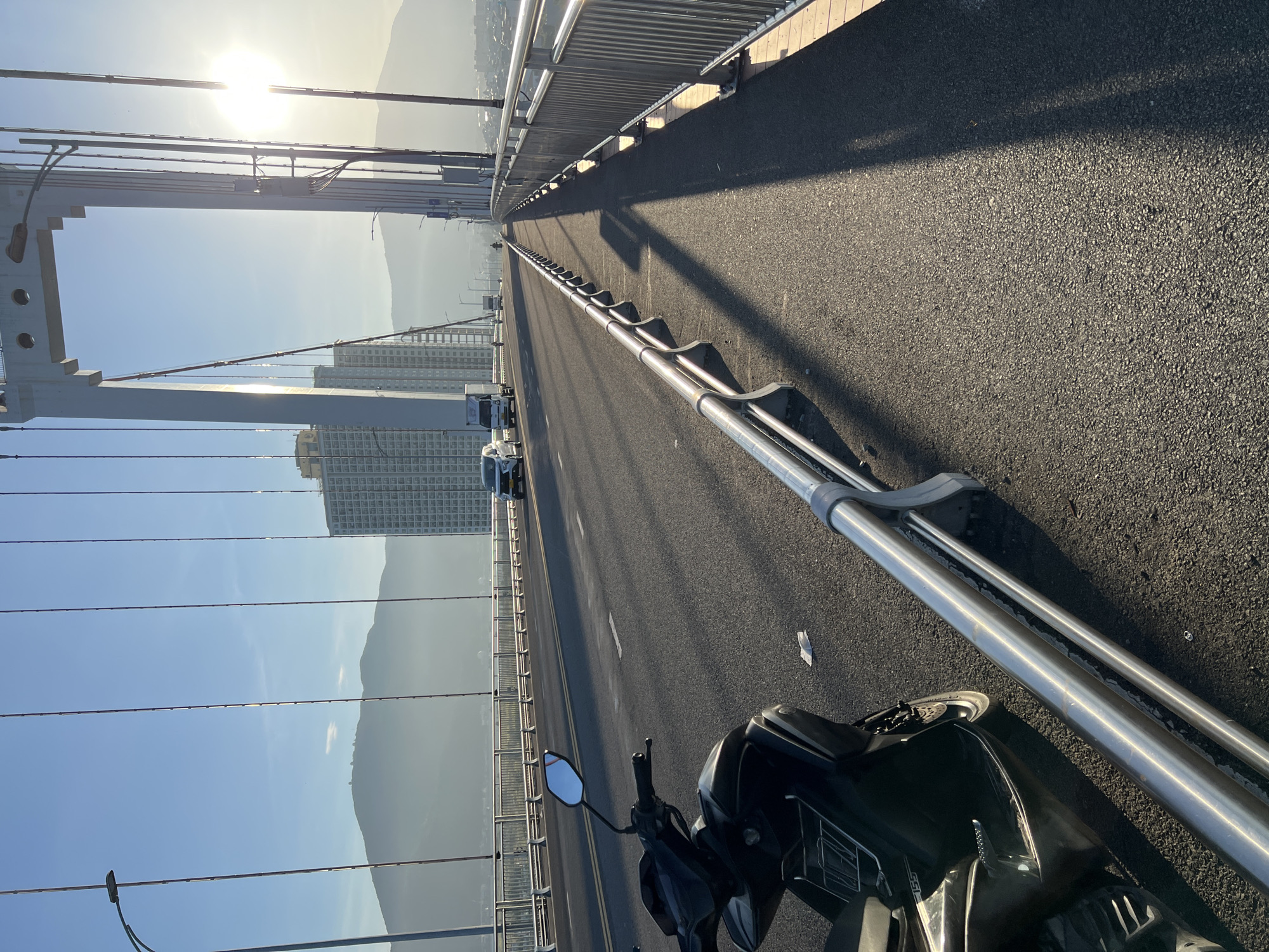 The image captures a view from a pedestrian walkway on a suspension bridge in Da Nang, Vietnam, with a motorcycle parked on the left. The sun is setting or rising, casting long shadows across the asphalt road and walkway. The bridge's structure, including suspension cables and a large white pillar, is visible. In the background, a tall building stands out against a hazy mountainous landscape. Cars and a truck are seen driving on the bridge. The sky is clear blue with a bright sun.