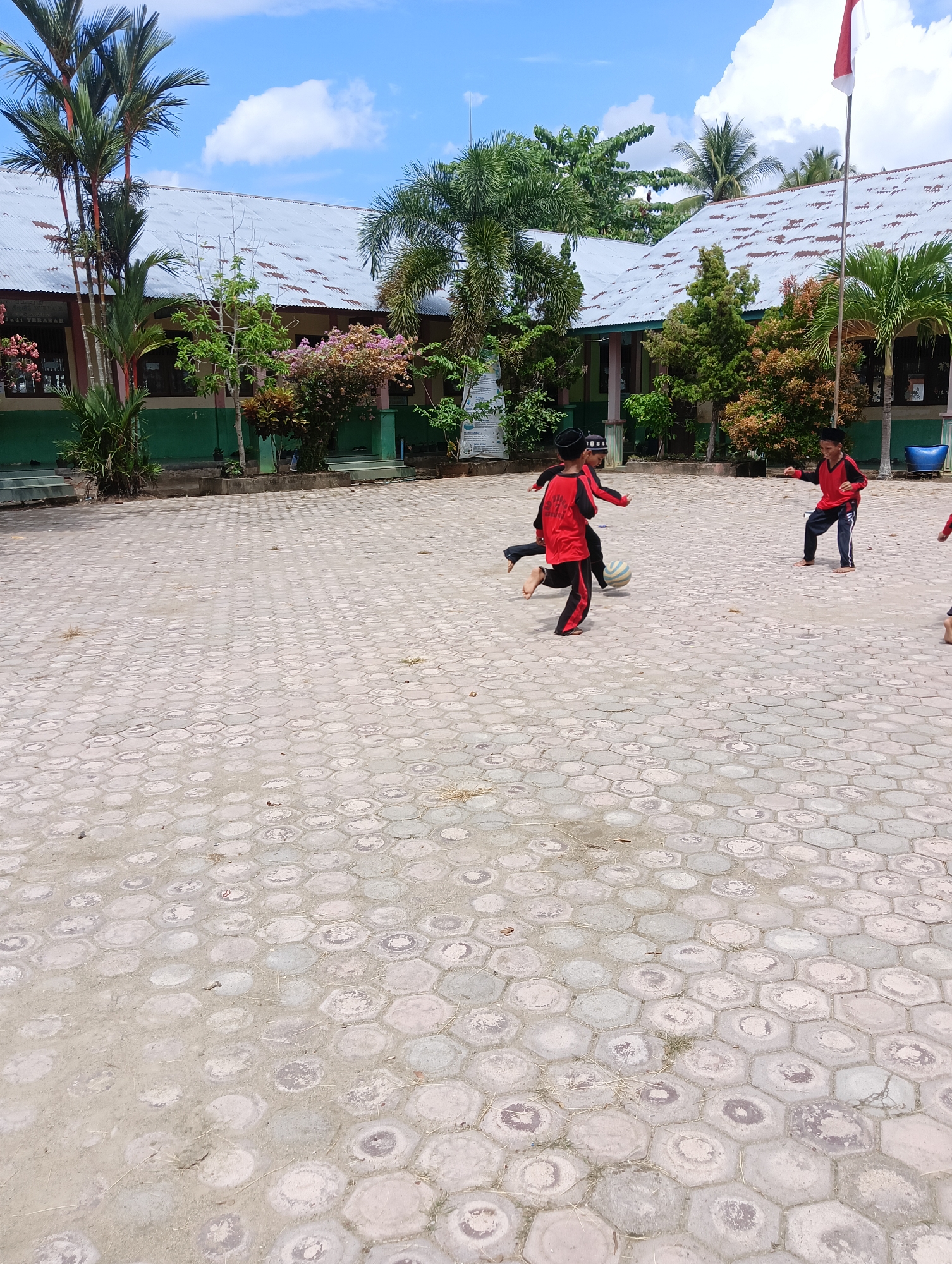 Four young boys in red shirts and black pants are playing soccer on a paved courtyard in front of a school. The school building has a metal roof and is surrounded by trees and greenery. The sky is blue with some clouds. The boys are actively engaged in the game, with one boy kicking the ball, another running towards it, and two others observing or participating. The ground is paved with hexagonal concrete tiles.