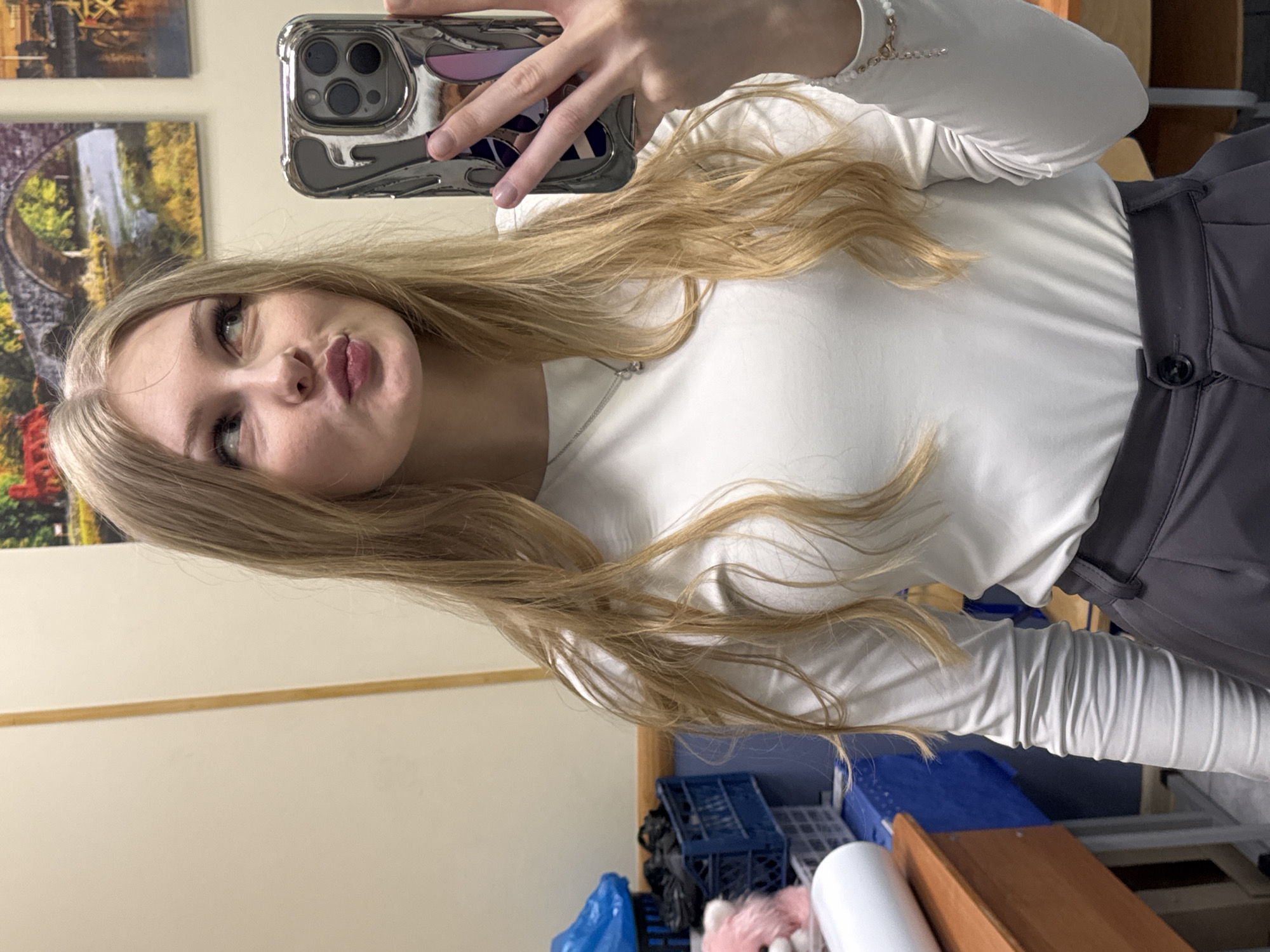 A blond-haired woman in a white long-sleeved shirt and grey pants is taking a selfie in an indoor setting. She is holding a metallic phone in her right hand. The background includes artwork on a wall, possibly depicting landscape scenes. Visible behind and below her are organizational items, potentially storage containers. The location is likely in Perm, Russia.