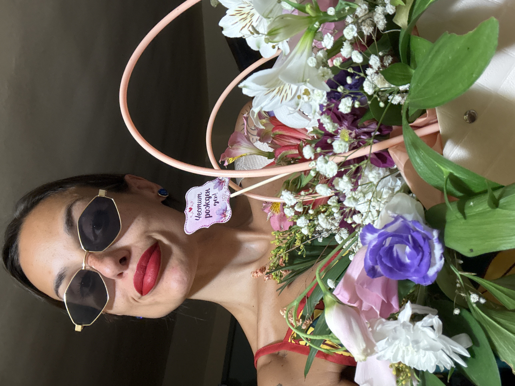 A woman with sunglasses and red lipstick holds a bouquet of flowers. A small sign with Cyrillic text reads "Честит рожден ден!" which translates to "Happy Birthday!". The woman is wearing a red strap top. The image appears to be taken indoors. There is no visible indication of the city of Pleven, Bulgaria.