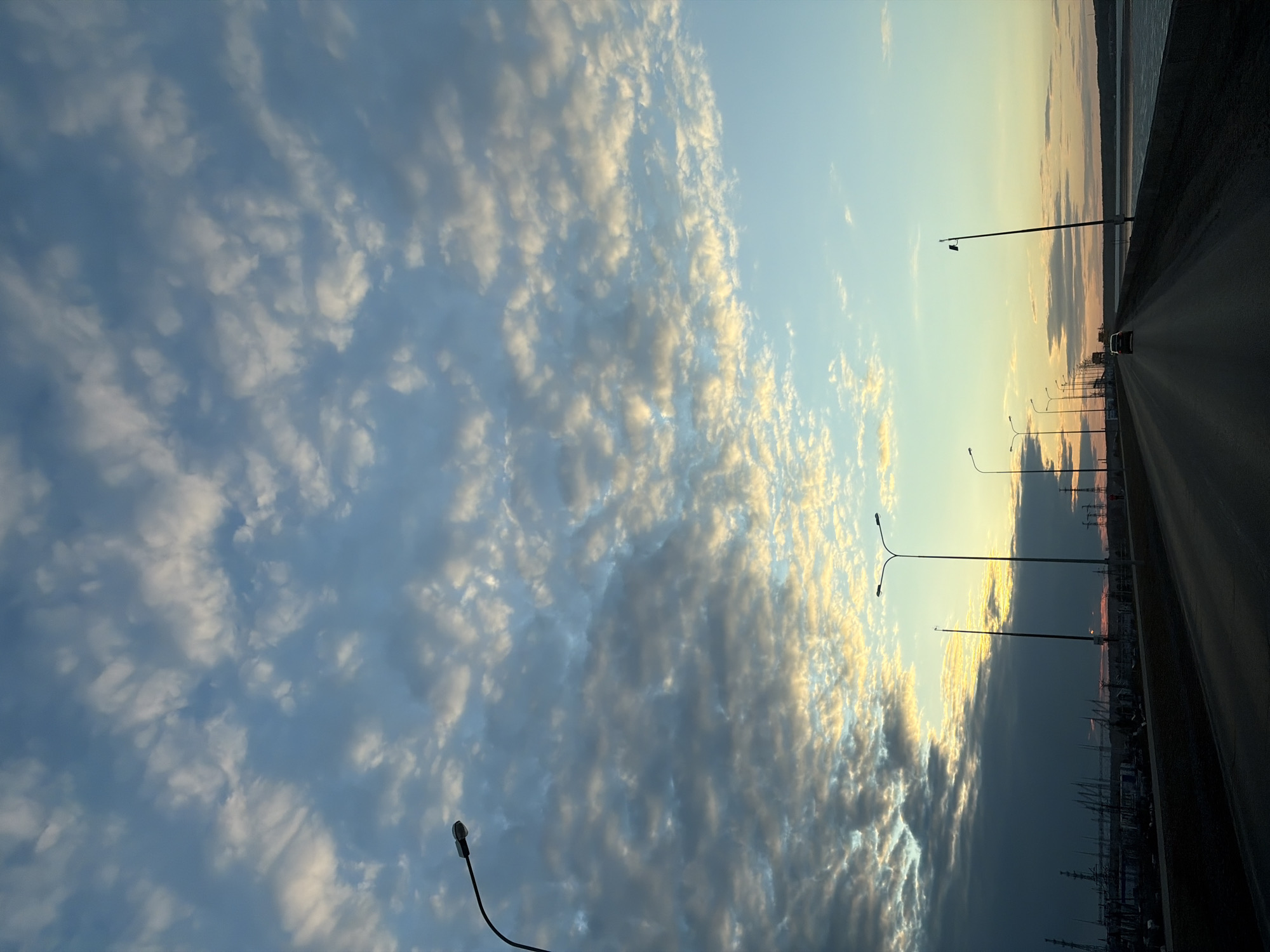 The image depicts a section of road leading into the city of Chaikovsky, Russia. The road, lit by evenly spaced street lights, stretches into the distance. In the distance, a vehicle is visible. To the side of the road, power lines and structures are situated. Above, a sky filled with textured clouds is visible. The sky is light blue with yellow highlights on the clouds near the horizon.