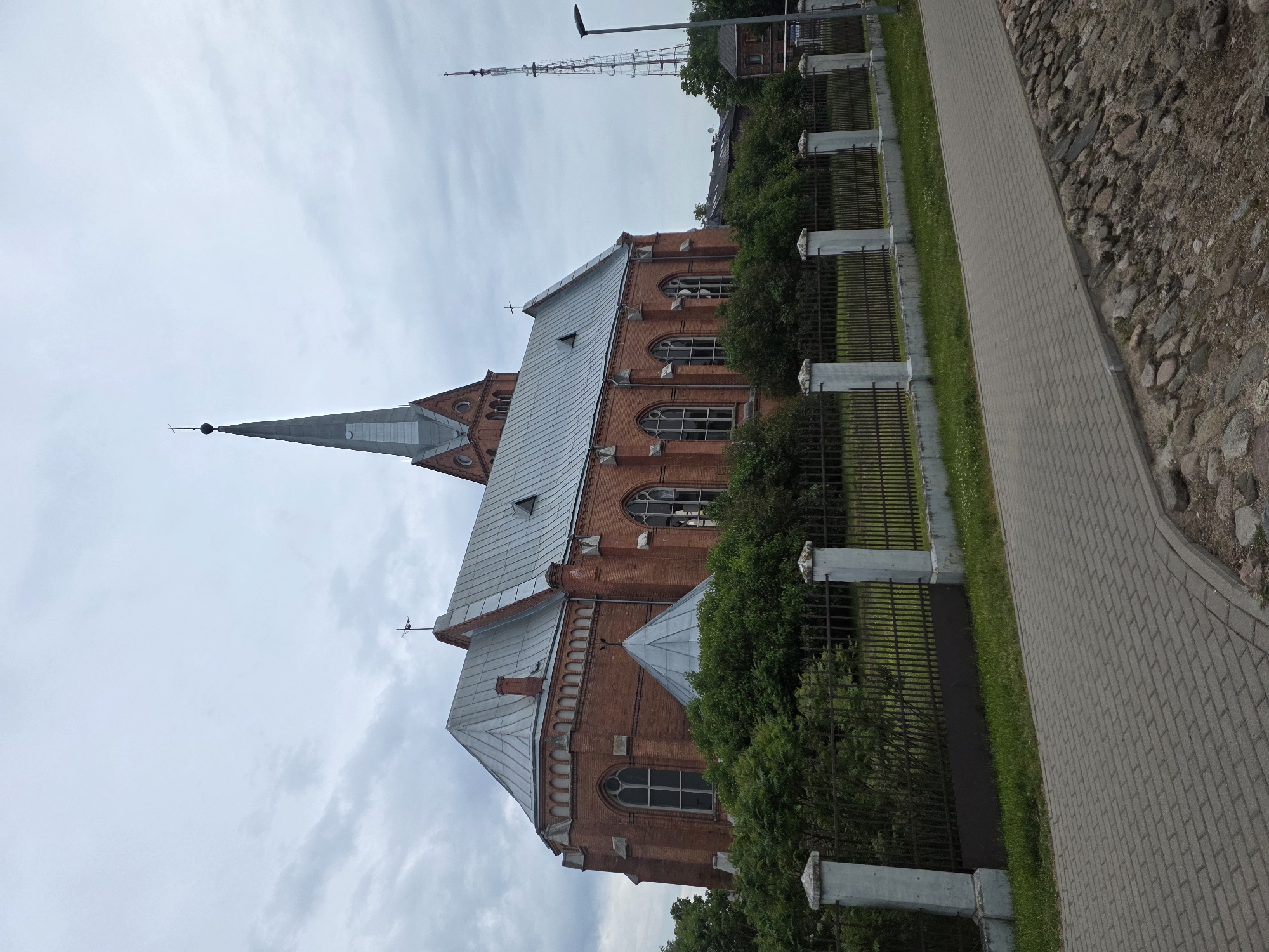 The image displays a red brick church with a prominent grey metallic roof and a tall, slender spire, situated in the city of Unknown, Unknown. The spire is topped with an orb and a cross, and additional crosses are visible on the rooflines. The church features arched, multi-pane windows and various roof structures, including small triangular dormers and a conical metallic roof on the side. A dark metal fence with light-colored concrete posts runs in front of the church, bordered by a strip of green grass. A grey paver sidewalk extends parallel to the fence, adjacent to an area of rough cobblestones. In the distance to the right, a tall, lattice communication tower and a black streetlight stand against an overcast sky. Several small, dark-roofed buildings are partially visible behind the church. No individuals are present in the scene.
