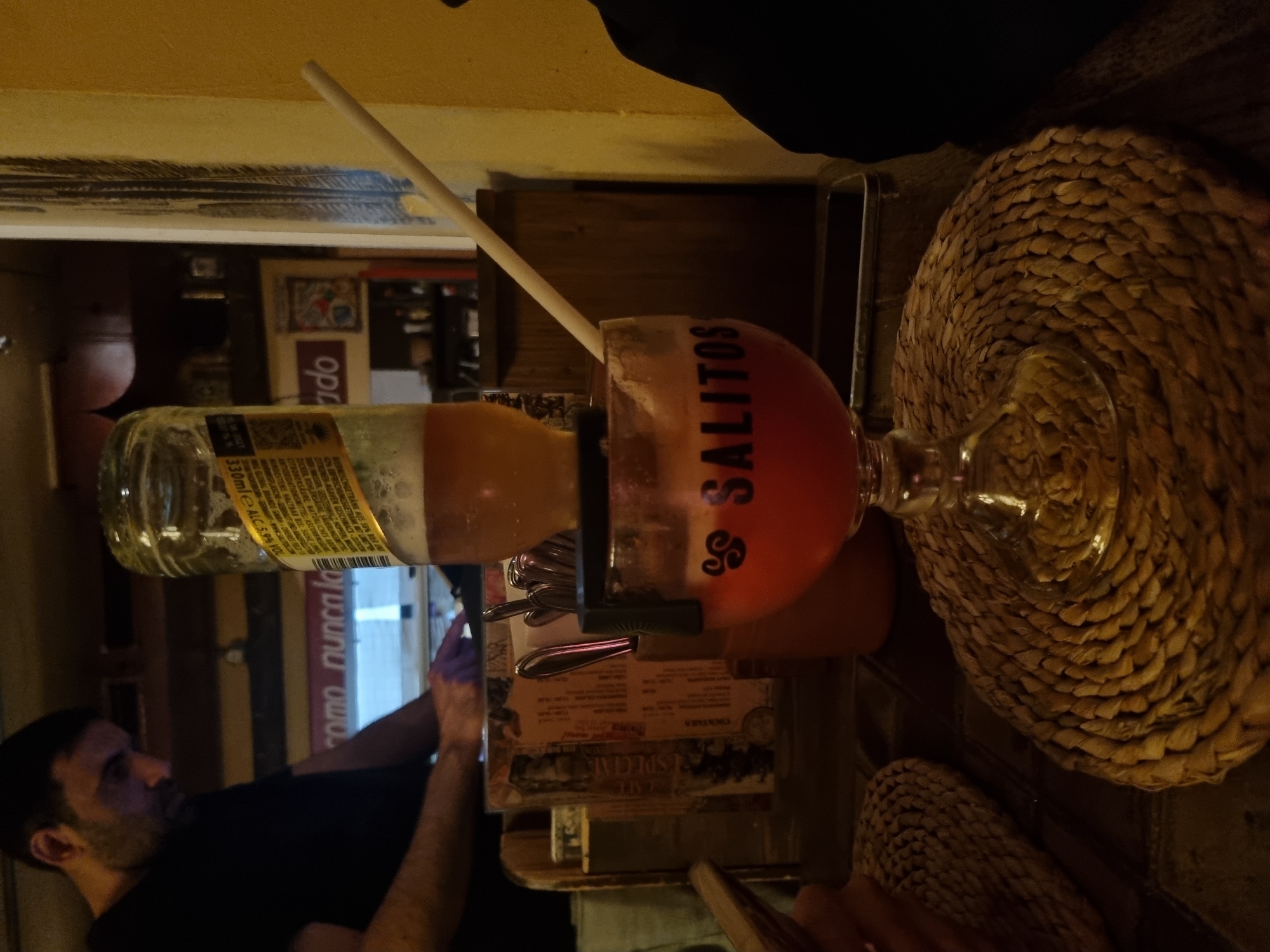 The image shows a "Salitos" cocktail, a blend of tequila, beer, and fruit juice, presented in a glass with an inverted beer bottle inserted into it. A long straw is also in the glass. The cocktail is placed on a woven placemat. There is a person visible in the background wearing a black shirt and reaching towards the bar. In the background, there is text that looks like "como nunca la", implying that the place is called "como nunca" - a bar in Saarbrücken, Germany. There's also a cocktail menu and a cutlery holder visible.