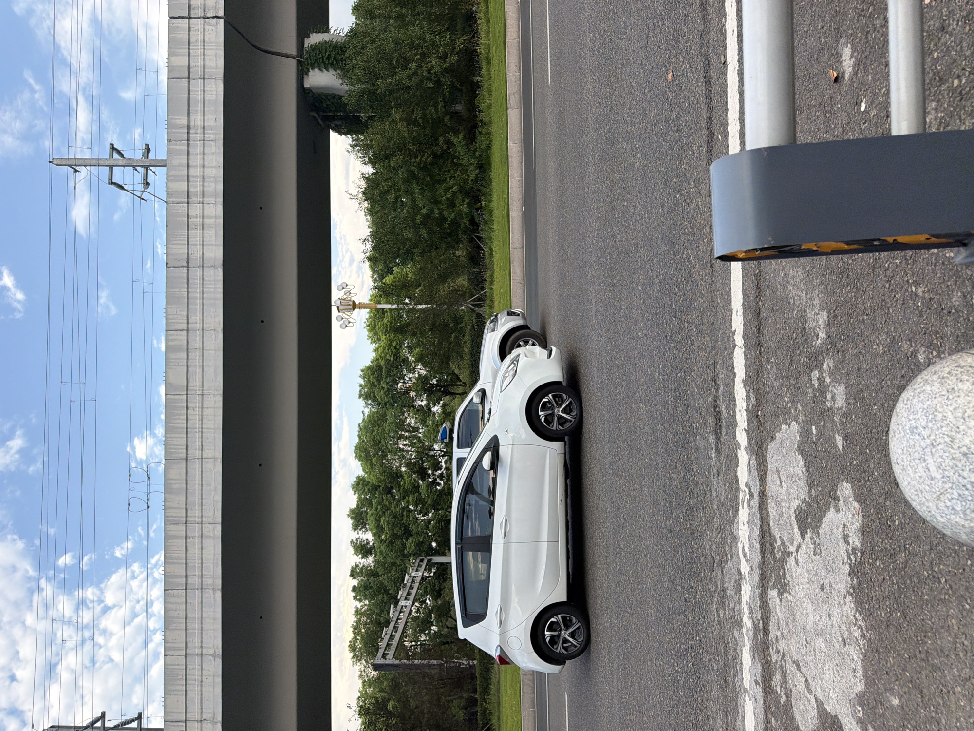 The image depicts a street scene in Jiangjin, China, likely on a moderately overcast day. In the foreground, the asphalt road surface is visible with white lane markings. A gray barrier with yellow accents is positioned at the lower right corner of the frame. Further into the mid-ground, a white hatchback car is traveling on the road, followed by a silver SUV. Behind the vehicles, a line of lush green trees forms a backdrop. Above the trees, a large concrete overpass stretches across the frame, supported by columns, with power lines and a utility pole visible against a blue sky with scattered white clouds. The overpass appears to be part of a transportation infrastructure, potentially a highway or railway. A decorative lamppost with multiple lights is also visible amongst the trees on the left side of the frame. The overall impression is of urban infrastructure interacting with the natural environment.