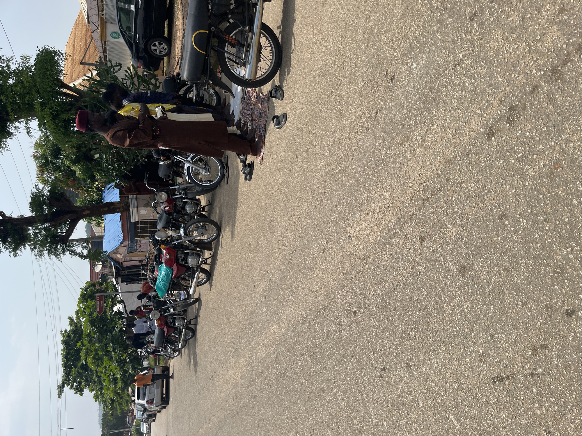 The image depicts a street scene in Abuja, Nigeria, with a row of parked motorcycles lined up along the side of a paved road. Several people are visible in the frame. In the foreground, a man wearing a brown robe and a red cap is standing, holding what appears to be prayer beads. Nearby, another person is partially visible, dressed in a yellow and blue shirt. Further down the line of motorcycles, other individuals can be seen, some standing and others seemingly tending to the motorcycles.
The scene suggests a busy street, possibly a marketplace or a transportation hub, where motorcycles are a common mode of transport. The time of day appears to be daytime, with bright sunlight casting shadows on the road, indicating fair weather. The presence of trees and some buildings in the background provides context for the urban setting. The general atmosphere seems to be one of everyday activity.
A red signpost with white lettering is visible on a pole amidst the trees, but the text is not clearly legible. The motorcycles are varied in color, including red, black, and a silver/white model. Some of the motorcycles have passengers or luggage on them. The road surface is asphalt with visible gravel. The overall impression is a snapshot of daily life in an African city, with a focus on transportation and the presence of people going about their business.