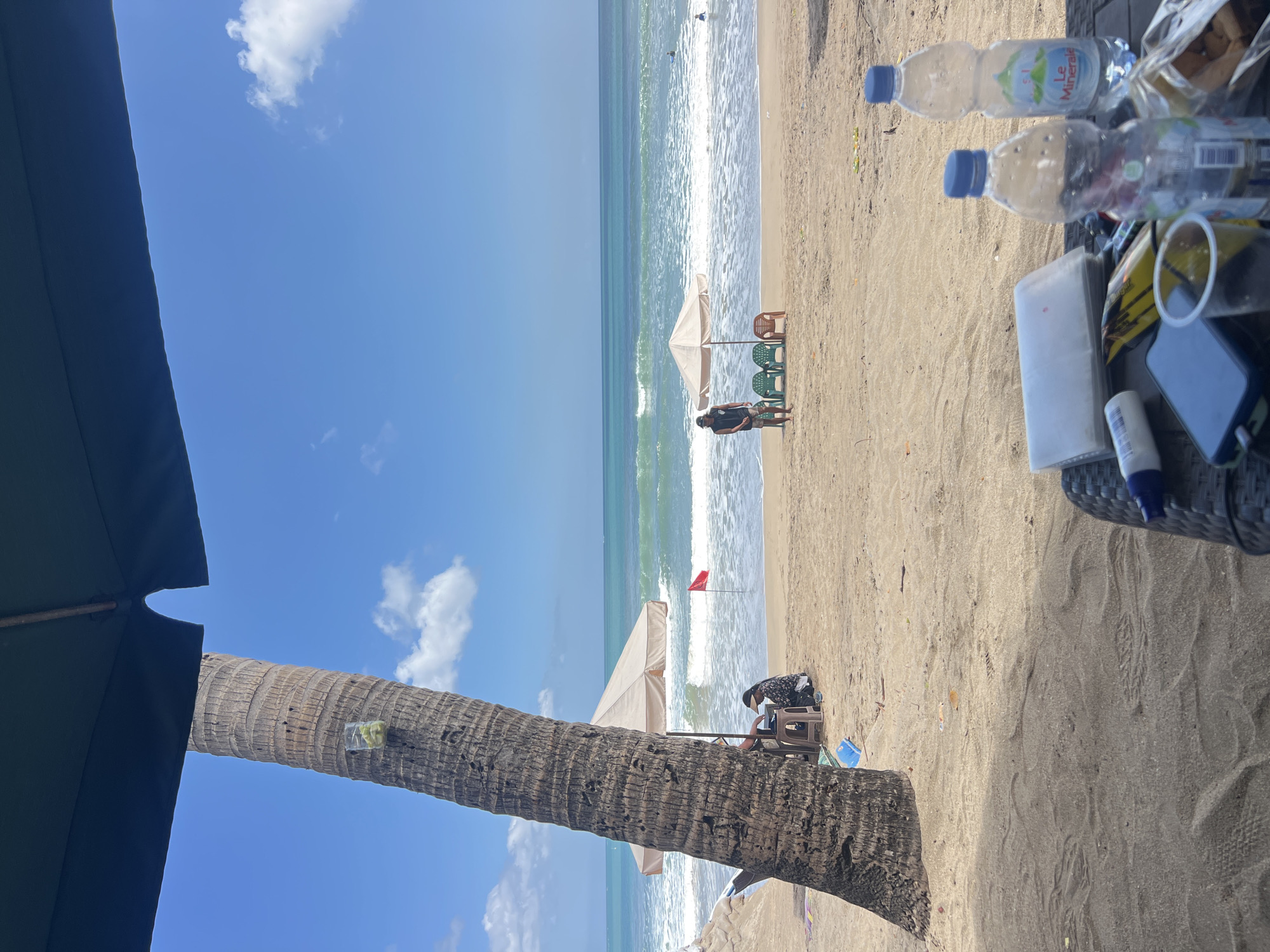 The image depicts a beach scene in Kuta, Indonesia, on a bright, sunny day. The foreground is occupied by a table laden with various items, including two plastic bottles of "Le Minerale" water, a phone, a small container, snacks, and a white rectangular object. A large palm tree trunk dominates the left side of the frame, with a clear plastic cup containing what appears to be green slices attached to it. In the midground, people are visible on the sandy beach. One person is seated under a tan umbrella near the water's edge, seemingly engaged with a table or object. Another individual stands near a set of beach chairs and a small umbrella. The turquoise ocean stretches to the horizon under a clear blue sky with scattered white clouds. Gentle waves break on the shore, and a few people can be seen in the water, possibly surfing or swimming. The overall atmosphere suggests a relaxed day at the beach.