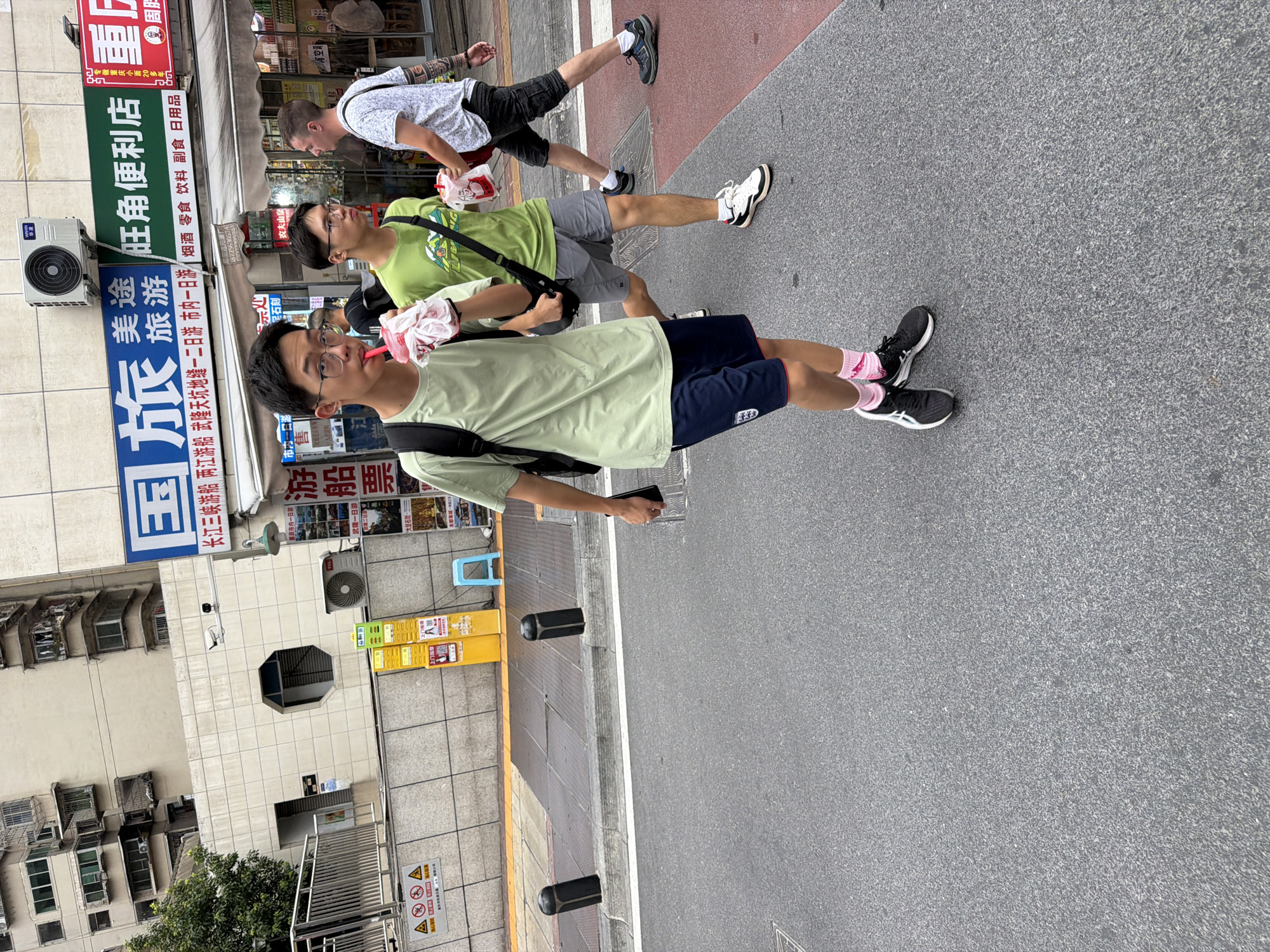 A medium shot captures three young men walking on a city street in Yuzhong District, China. The man in the foreground, wearing a light green t-shirt and dark blue shorts, is holding a pink cup with a straw and appears to be drinking from it. He has dark hair and glasses. To his right, another man in a green t-shirt and grey shorts is walking, also holding a pink cup. The third man, on the far right, is wearing a white patterned t-shirt and dark shorts and has tattoos on his arm. He is walking with his head turned to the left, possibly speaking to the man in the green t-shirt. The background features a multi-story building with visible air conditioning units and a street sign with Chinese characters. The ground is asphalt, with yellow and grey bollards visible on the sidewalk. The overall scene depicts a casual street interaction in an urban environment.