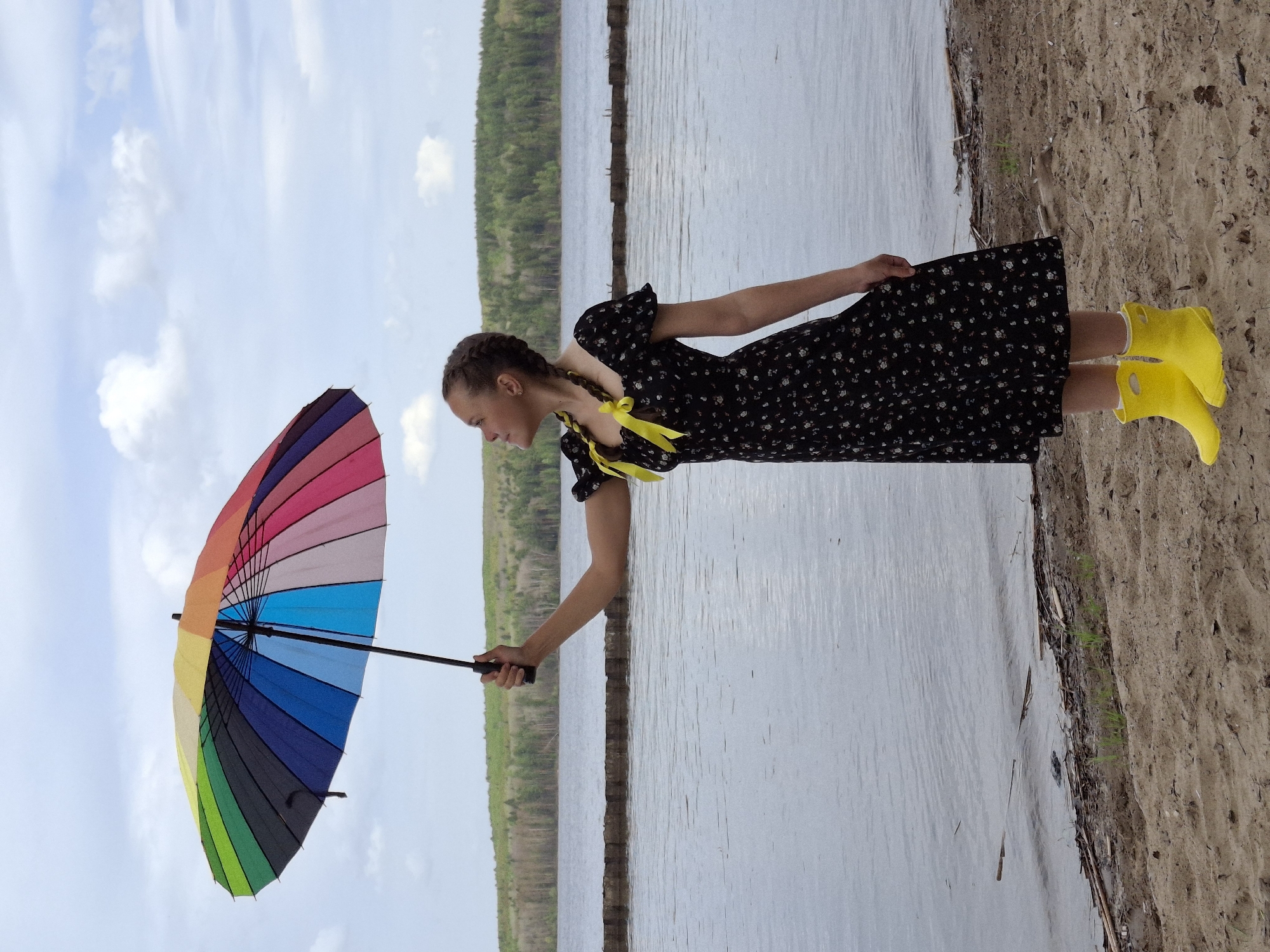 A young woman stands on a sandy shore near a body of water. She is holding a rainbow-colored umbrella. She wears a dark, floral-patterned dress, yellow bows in her braided hair, and yellow rain boots. In the background are trees and a cloudy sky. The location is Chaikovsky, Russia.