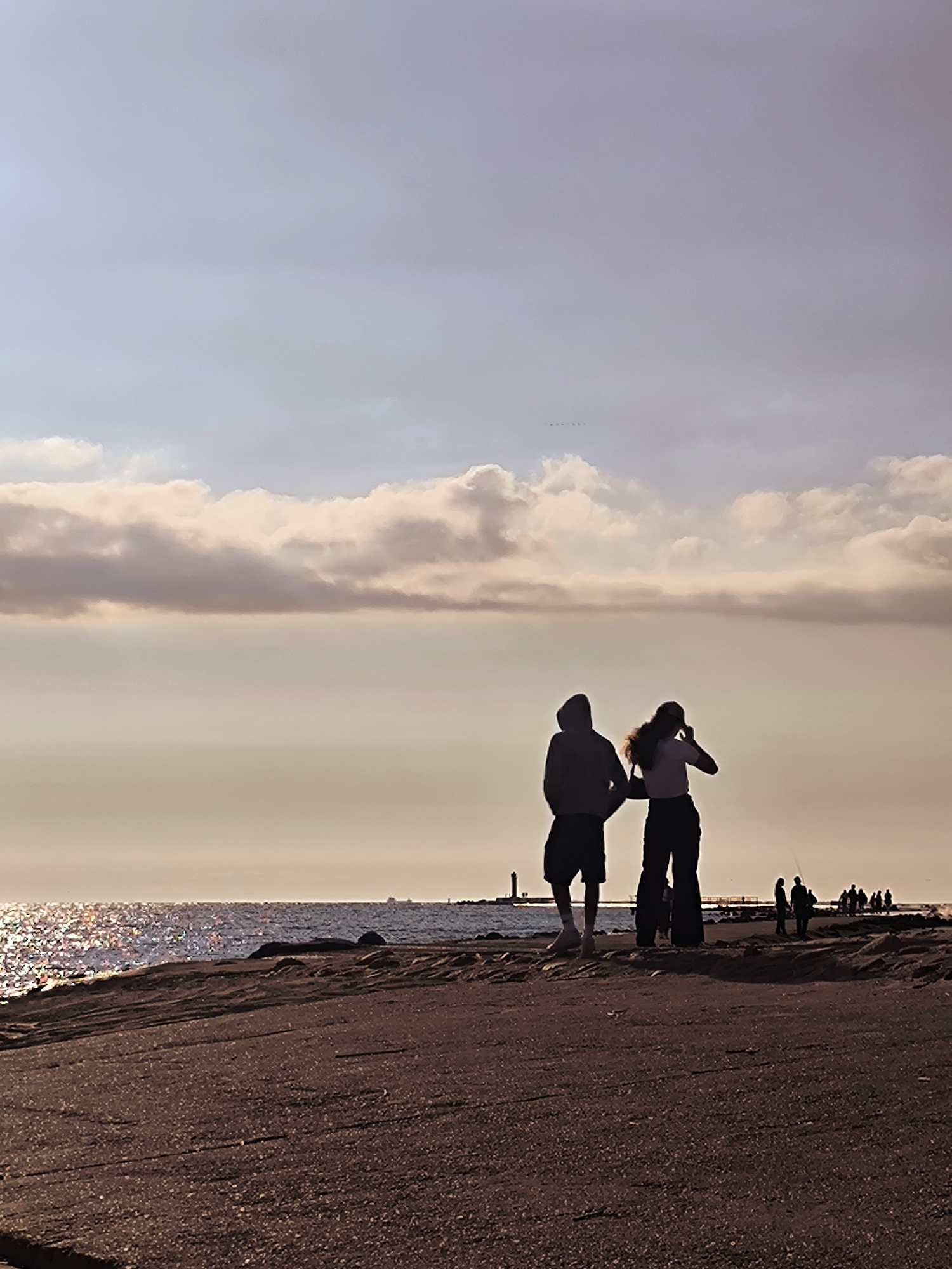 The image shows a silhouetted pair of people walking on a paved walkway at sunset, overlooking a body of water that reflects the warm hues of the sky. The walkway is textured with small stones and appears to be part of a pier or promenade. The water is calm, with a distant lighthouse visible on the horizon. Further in the background, a group of smaller silhouetted figures are gathered. The sky is filled with soft, illuminated clouds. The scene evokes a sense of tranquility and end-of-day reflection. This location is likely the Jurmala beach promenade in Riga, Latvia, a popular spot for evening strolls and viewing sunsets over the Baltic Sea.