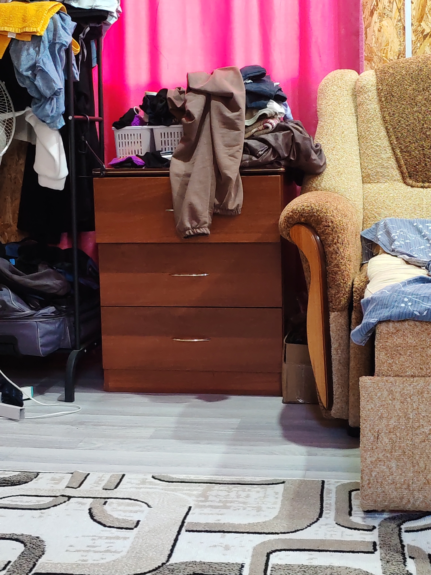 This is a medium-shot, eye-level interior photograph of a cluttered room. In the center, a wooden dresser with three drawers is visible, with a gray garment draped over the top drawer. To the left of the dresser, a black metal clothing rack holds several items of clothing, including a yellow towel at the top, blue shirts, and a white garment hanging down. A white laundry basket containing dark-colored clothing sits on top of the dresser. To the right of the dresser is a portion of a beige, textured armchair with a brown and yellow patterned throw blanket. A light pink curtain hangs in the background. The floor is covered with a light-colored laminate, and a rug with a geometric pattern is visible in the foreground. The scene is set in a room in Kuyeda, Russia.